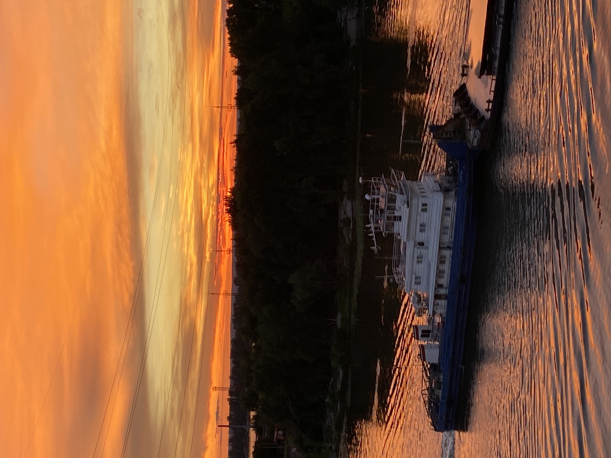 A barge is being pushed down a river in Novokuybyshevsk, Russia, at sunset. The sky is a vibrant orange and yellow, with some clouds. The trees on the riverbank are dark silhouettes against the colorful sky. The water is reflecting the colors of the sunset, creating a shimmering effect. There are power lines in the background.