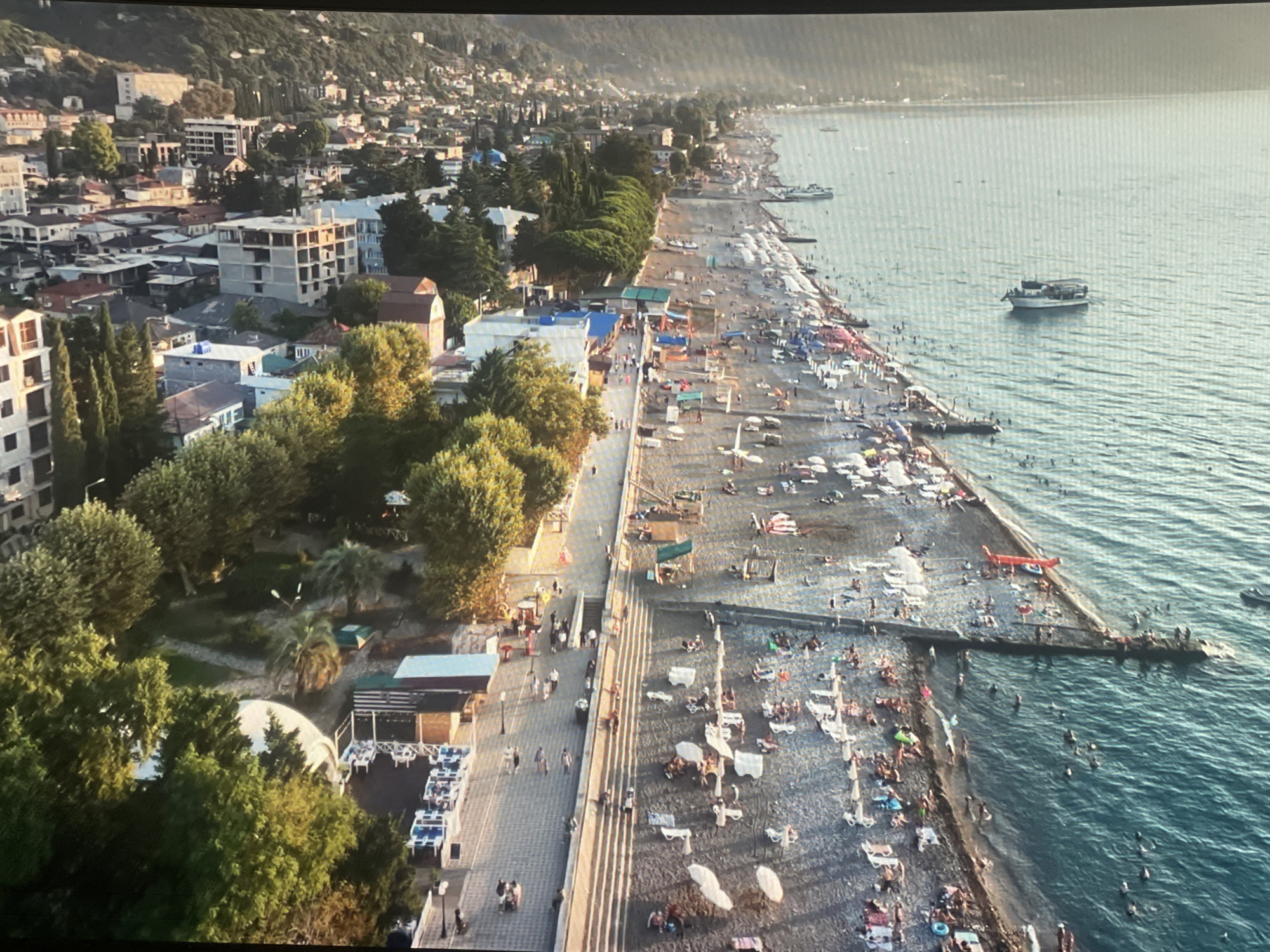 This aerial view captures a bustling seaside resort town, likely Sokhumi, Abkhazia, during a sunny day. A long, pebble beach stretches along the coastline, populated by numerous sunbathers relaxing on lounge chairs beneath a scattering of umbrellas. People are also seen wading and swimming in the clear, turquoise waters of the sea. Adjacent to the beach, a paved promenade is lined with trees and small buildings, possibly shops or cafes. A prominent pier extends into the water, where a boat is docked, and people are gathered. In the background, a dense cluster of buildings, ranging from multi-story structures to smaller houses, ascends a green hillside, indicative of the town's urban development. Several boats are visible in the sea, some near the shore and others further out. The overall scene depicts a vibrant summer day at a popular coastal destination.