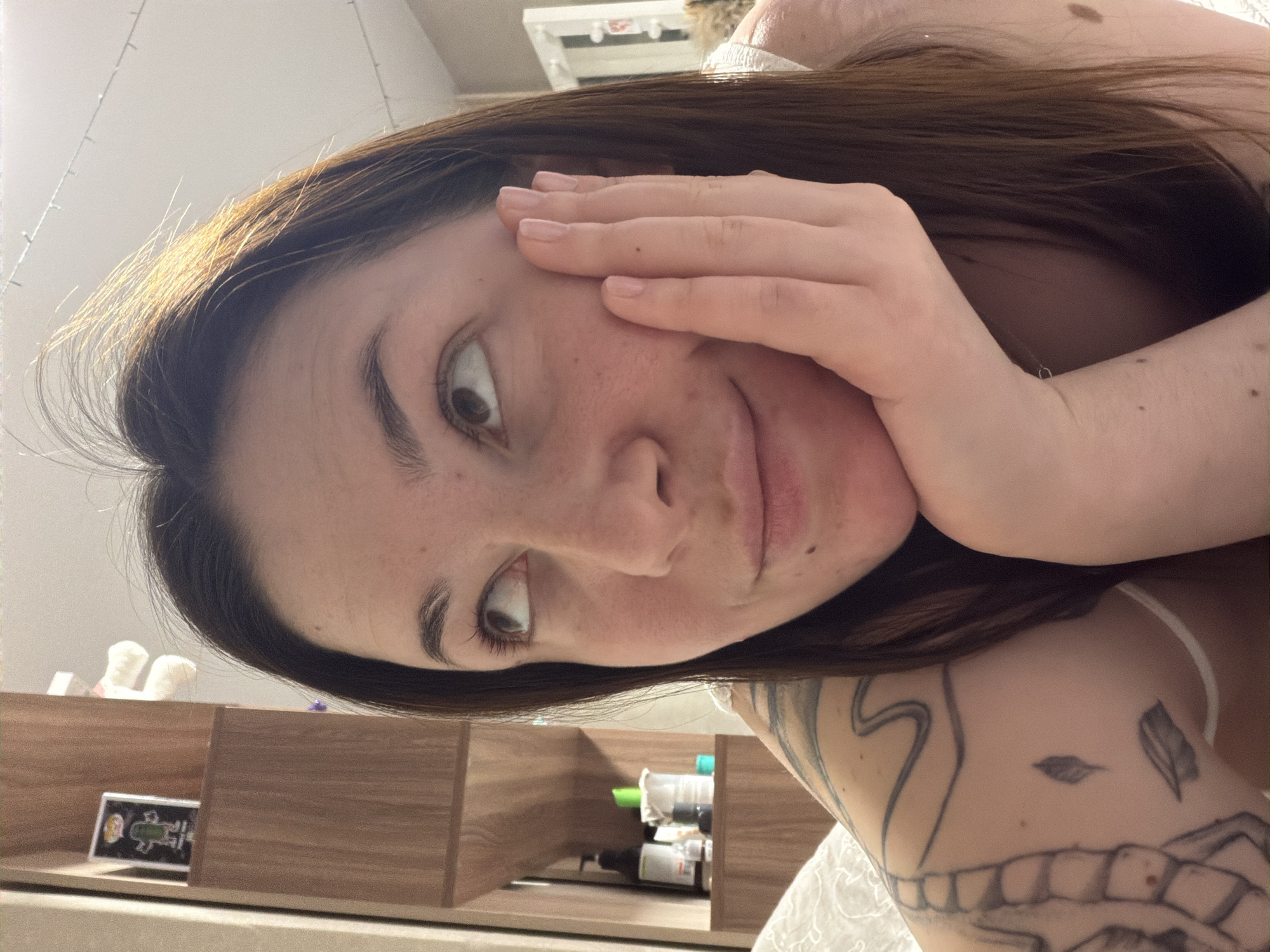 A young woman with brown hair and fair skin is looking at the camera with a slight smile. She is holding her hand to her face. She has a tattoo on her arm. In the background, there is a wooden shelf with various objects on it. There is no information in the image to suggest that the location is in the city of Chaikovsky, Russia.