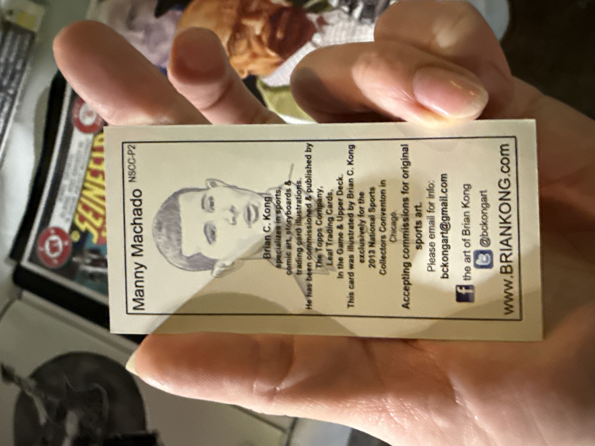 A hand holds a business card for Brian C. Kong, an artist specializing in sports and comic art. The card features a black and white illustration of baseball player Manny Machado and includes Kong's contact information: email address, social media handles, and website. The card also states that it was exclusively illustrated for the 2013 National Sports Collectors Convention in Chicago.