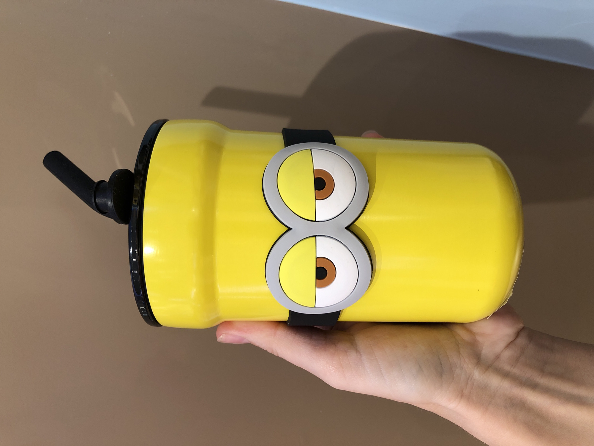 This media file shows a yellow Minion-themed reusable water bottle being held by a person's hand. The bottle is cylindrical and features two large, cartoonish eyes, a signature element of the Minions from the "Despicable Me" franchise. The eyes are depicted with grey circular frames, white irises, and brown pupils. A black band encircles the bottle, connecting the two eyes. A black straw extends from the lid of the bottle. The background consists of a gradient of warm brown and a light blue surface, suggesting an indoor setting or an area with diffused lighting. The hand holding the bottle appears to be resting on a neutral-colored surface. The overall impression is of a playful and casual object being presented. There is no visible text in the image. The setting's warm tones and lighting do not provide specific cues for time of day or weather.