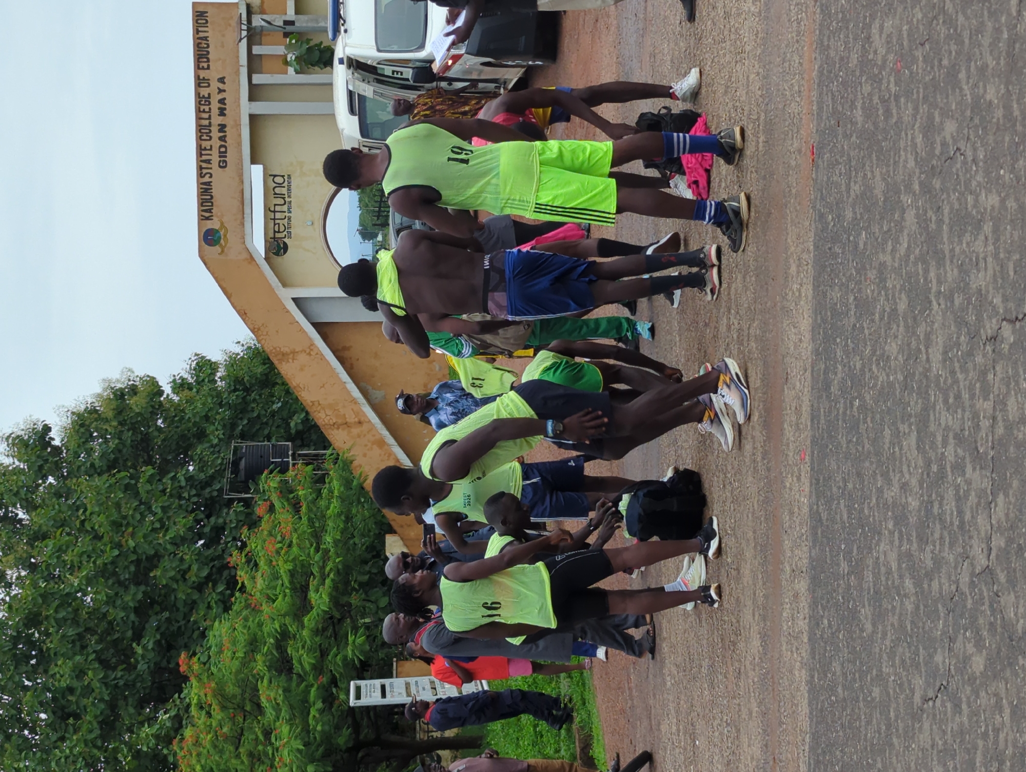 A group of young men are gathered near the entrance to the Kaduna State College of Education in Gidan Waya, Nigeria. They are wearing sports attire, including neon green vests with numbers, shorts, and athletic shoes. Some are bare-chested. They appear to be engaged in some sort of training or exercise activity. Some people are watching and recording the scene with mobile phones. A vehicle is parked in the background, and the college sign is visible.