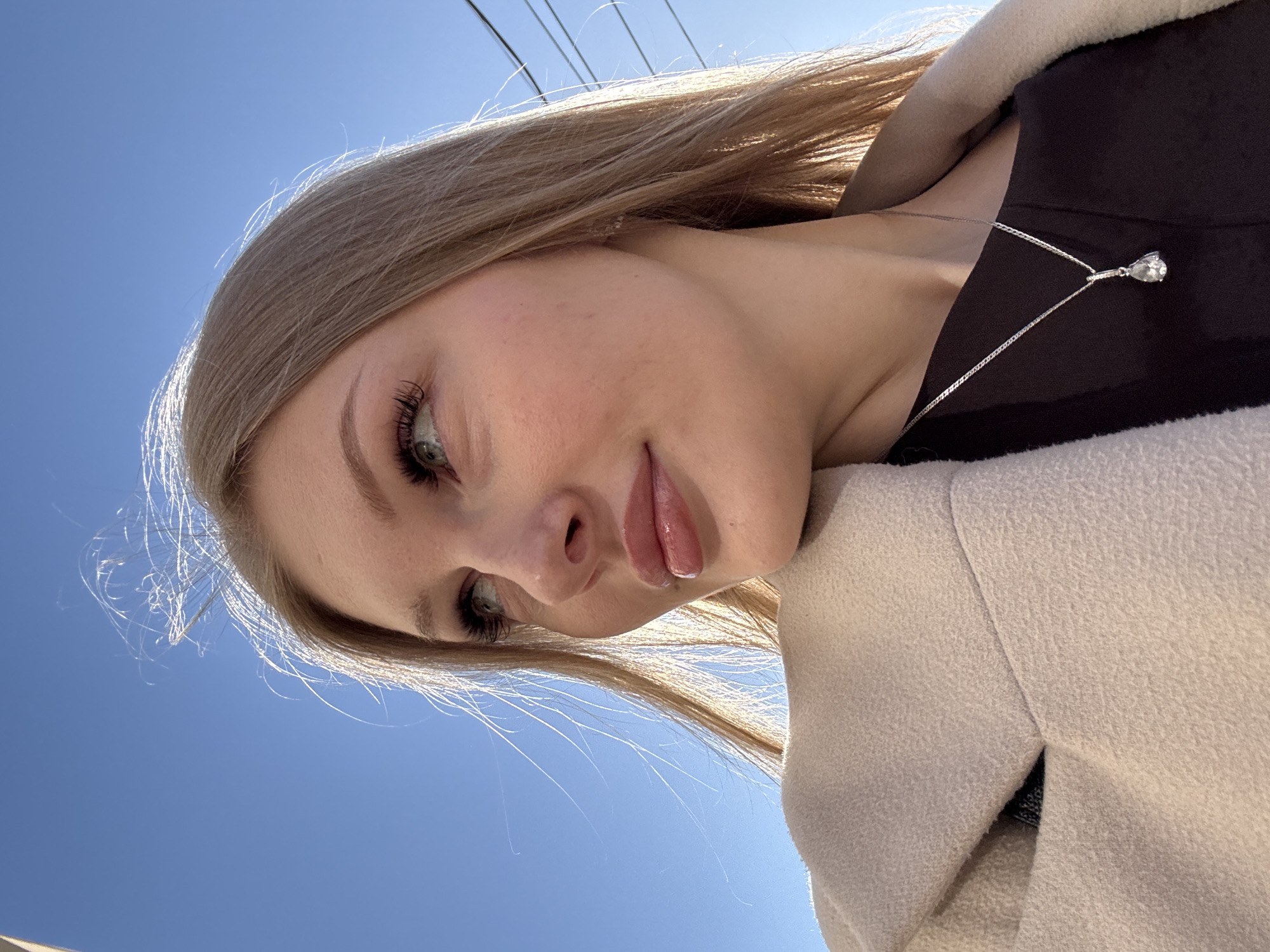 The image depicts a young woman with light blonde hair, wearing a black top and a light beige coat. A silver necklace with a teardrop-shaped pendant is visible. The woman is looking down and to the left. The background consists of a clear blue sky and power lines. It is not possible to determine the specific location within Perm, Russia from the image.