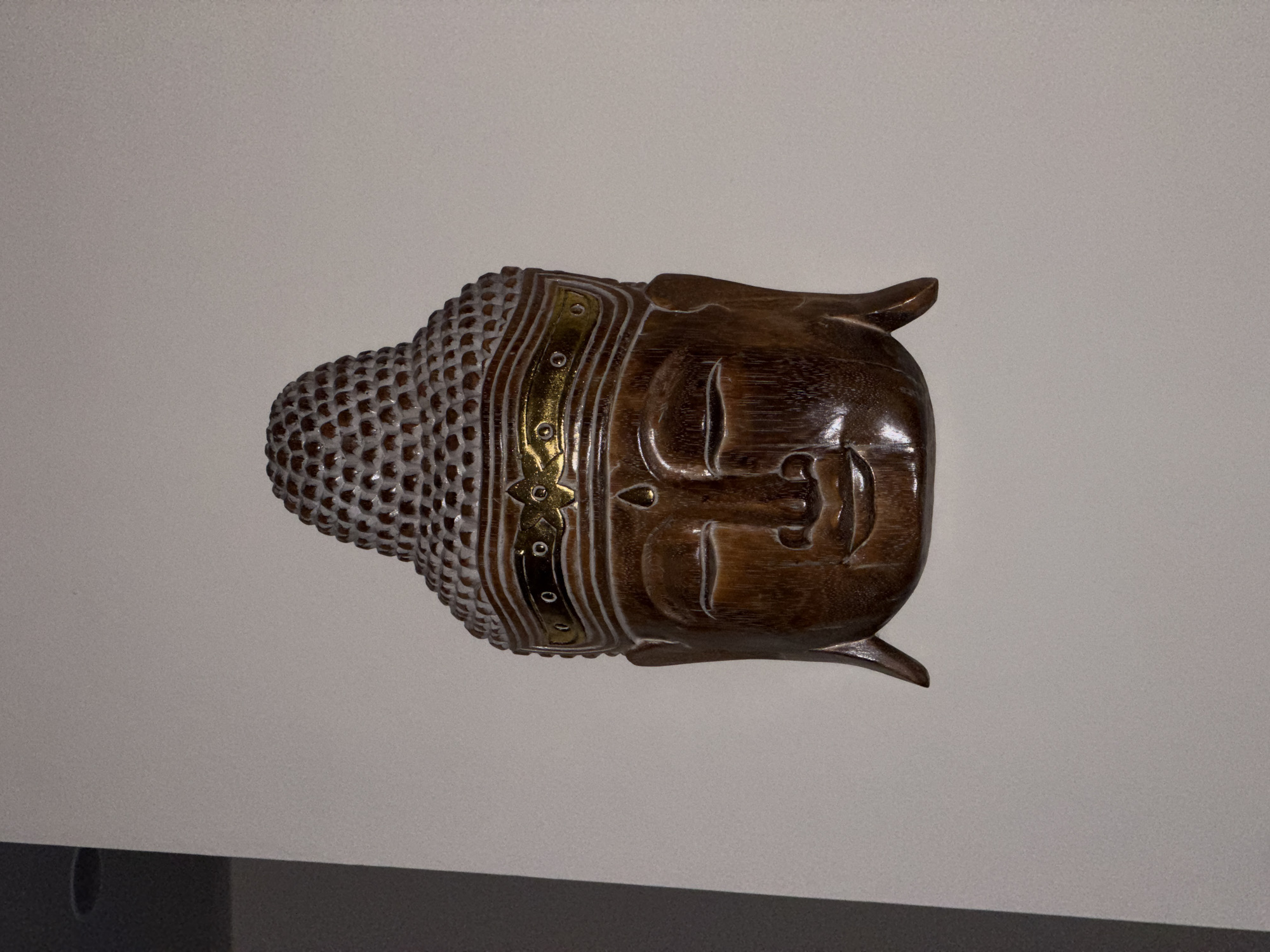 A wooden Buddha head sculpture is mounted on a light gray wall. The sculpture features a textured crown with small circular patterns, a decorative band with gold and white accents, and a smooth, stylized face with closed eyes and a serene expression. The overall color of the sculpture is a dark, warm brown. A dark gray surface is visible in the lower left corner of the image. No people are present. The location is Kalibukbuk, Indonesia.