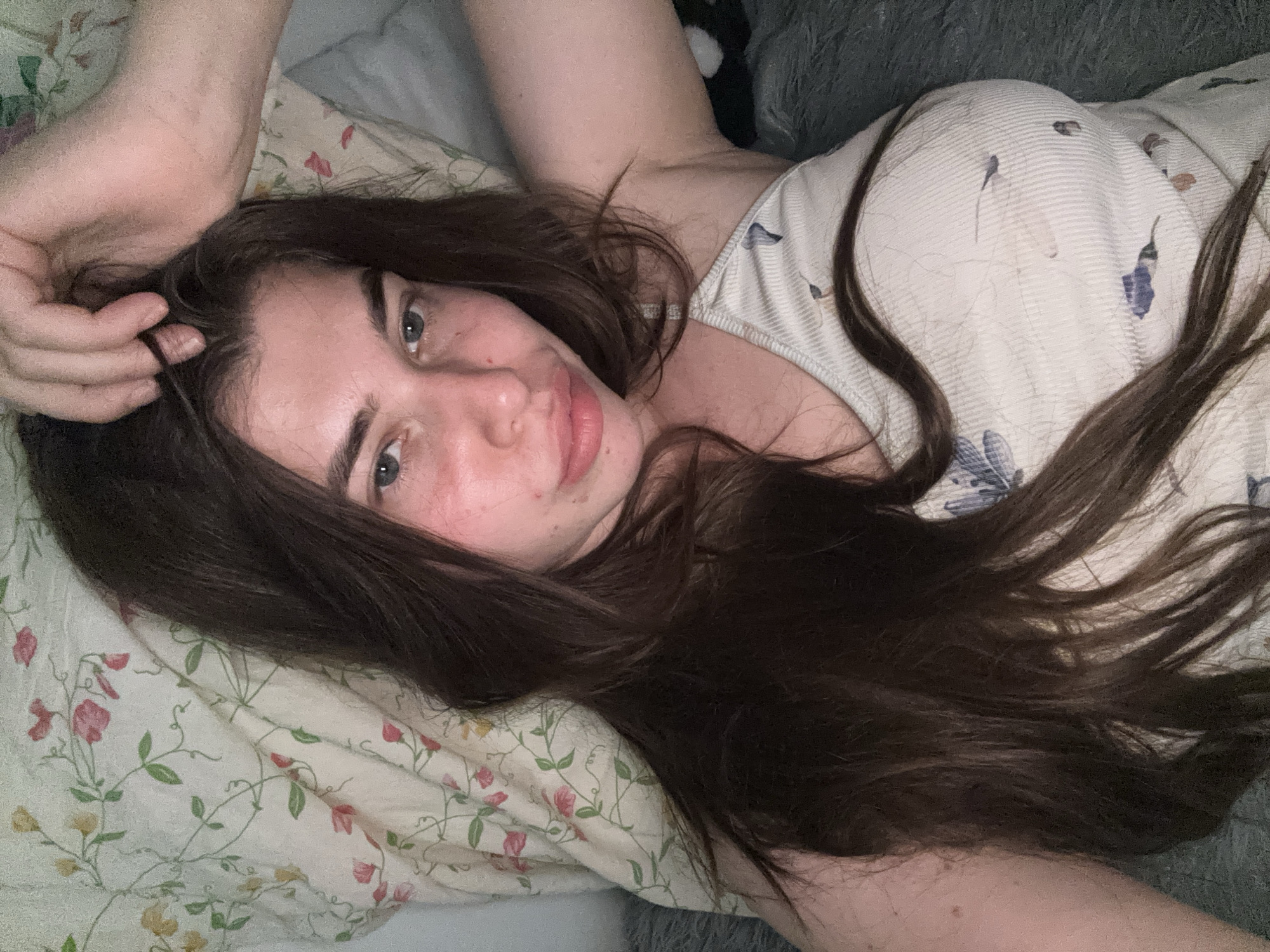 A young woman with long, dark brown hair and fair skin lies on a bed covered with a floral-patterned sheet. Her head is propped up on the pillow, and her left hand is resting on top of her head, touching her hair. She is looking directly at the camera with a neutral expression. She is wearing a light-colored ribbed tank top with a blue and white floral pattern. The background shows a pillow with a pink and green floral print. The overall scene suggests a casual, private setting, possibly a bedroom. The image does not provide any specific information about the location in the city of Мещёры, Russia.