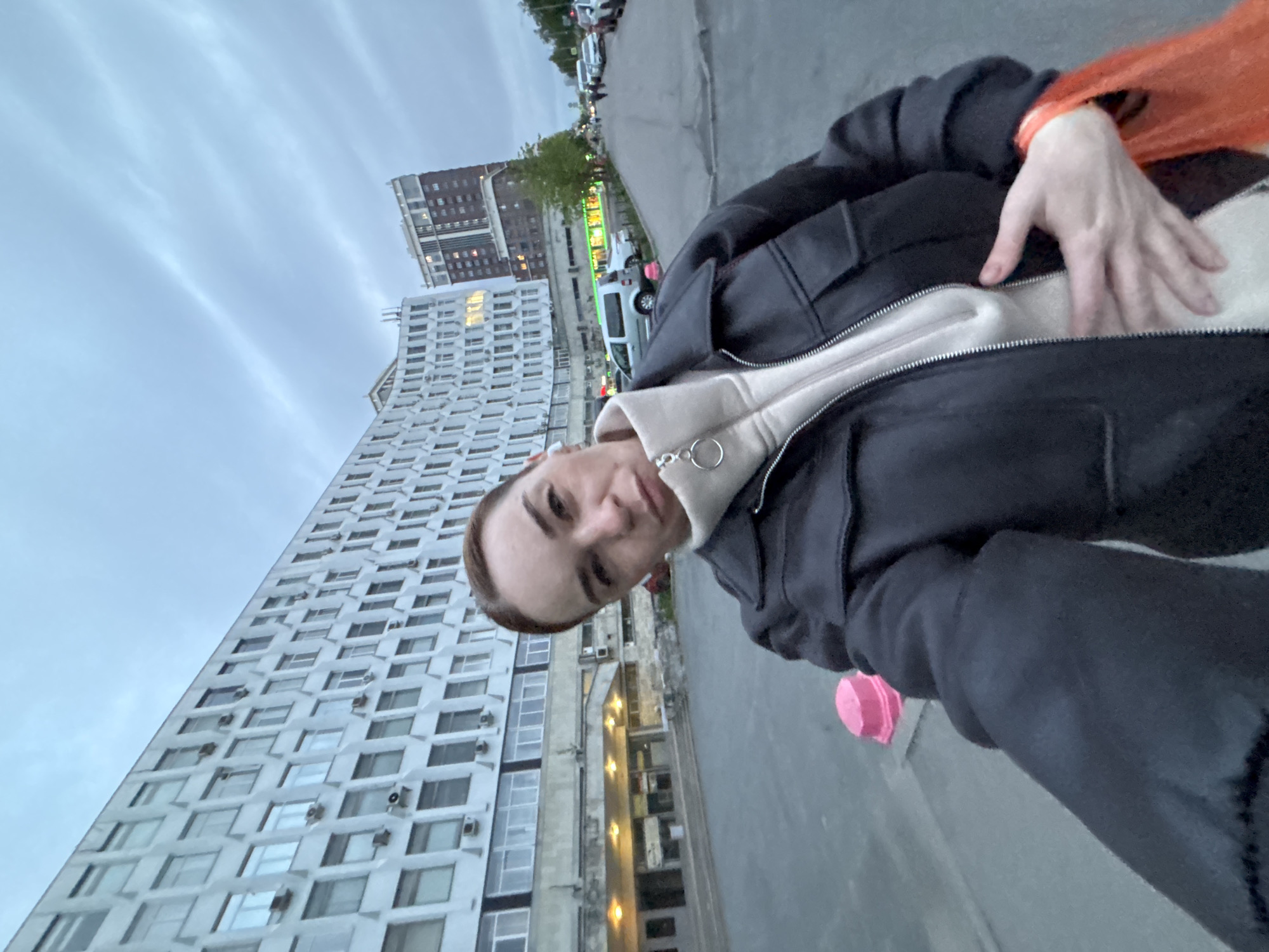 A selfie shows a person outdoors in an urban setting. The person is wearing a black jacket over a light-colored hoodie. In the background is a tall, light-colored building with many windows and air conditioning units. Another building with a darker facade can be seen further back. A street with parked cars is visible, and the sky is overcast. The person is holding the camera at an angle, resulting in a tilted perspective of the buildings and street.