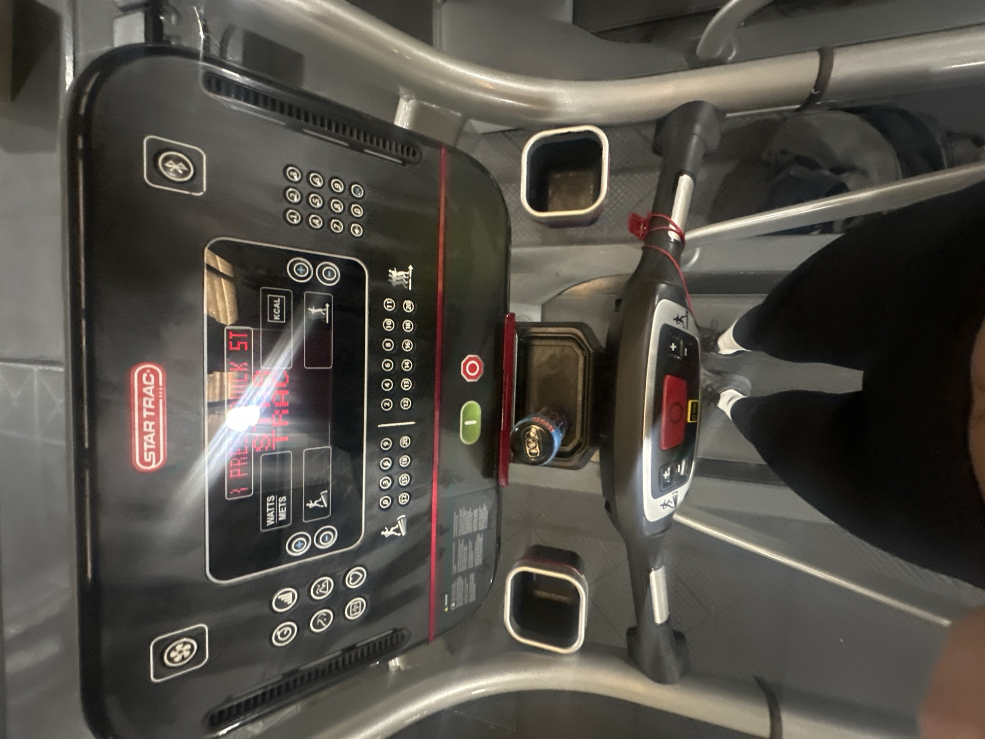 The image shows an overhead view of a Star Trac treadmill. The display shows the words "PRE" and some blurred numbers, presumably indicating a pre-set program and time. A person wearing black leggings and white socks stands on the treadmill. A red elastic band wraps around the treadmill handle. There is a can of soda in the console cup holder. The environment suggests an indoor gym setting. The location is Tonbridge and Malling, United Kingdom.