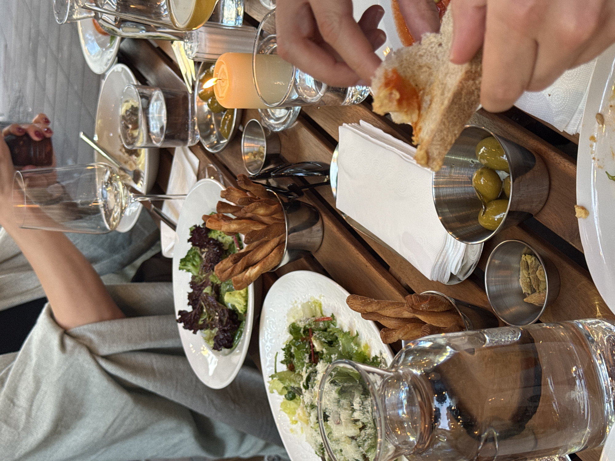 The image depicts an outdoor dining scene at a restaurant in Belgrade, Serbia. A table is laden with several dishes, including salads and breadsticks, alongside beverages and condiments. The ambiance suggests a pleasant meal shared among individuals, possibly friends or family, as evidenced by the presence of multiple plates and glasses. One person is seen in the background, partially obscured by a gray shawl, while another's hand is visible in the foreground, reaching for a piece of bread with topping. A lit candle adds a warm glow to the setting, and a carafe of water and wine glasses are also present. The focus is on the food and the convivial atmosphere of the dining experience.
