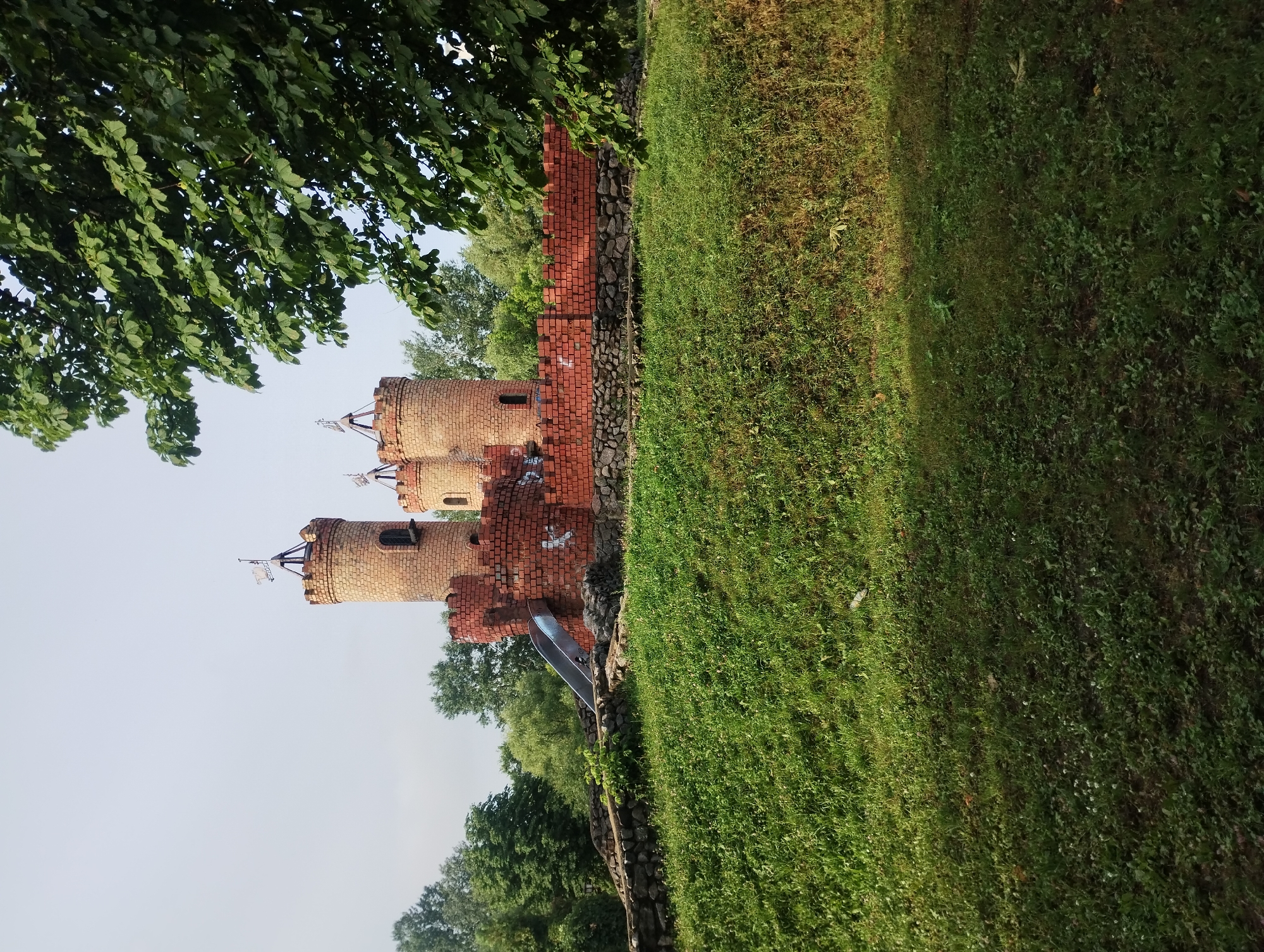 The image shows a medieval-style brick castle playground with two towers and a connecting wall, located in the city of Sumy, Ukraine. A silver slide is attached to the left tower. The castle is situated on a grassy hill, with trees and a clear sky in the background. The sun casts shadows on the grass, indicating it is daytime. There is no visible human presence in the image. The area appears to be a park or recreational space.