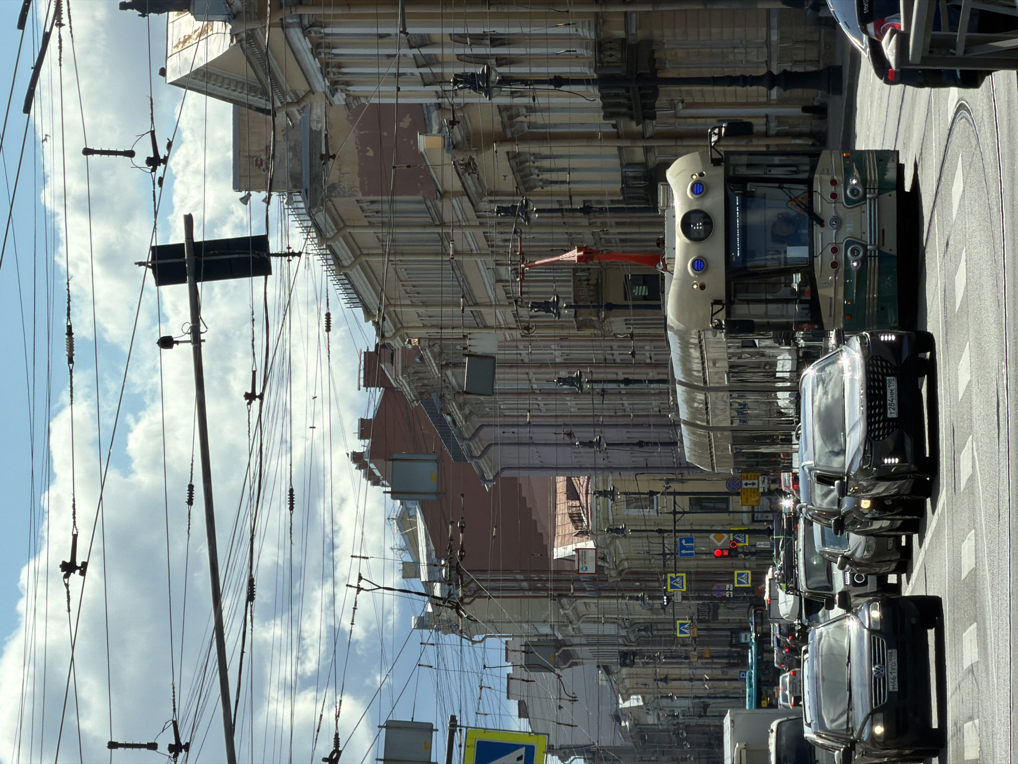 This is a vertical, eye-level, medium shot of a city street in Saint Petersburg, Russia, on a partly cloudy day. In the foreground, several cars are stopped at a red light, with a modern tram in the center lane. The tram is cream and green with blue lights on the front. In the background, classic Saint Petersburg architecture lines the street, with ornate facades and tall buildings. The sky is filled with dramatic clouds, and numerous overhead wires crisscross the scene, characteristic of a city with a well-developed public transportation system. The street is wet, possibly from recent rain. The overall impression is one of a bustling urban environment with a blend of historical and modern elements.