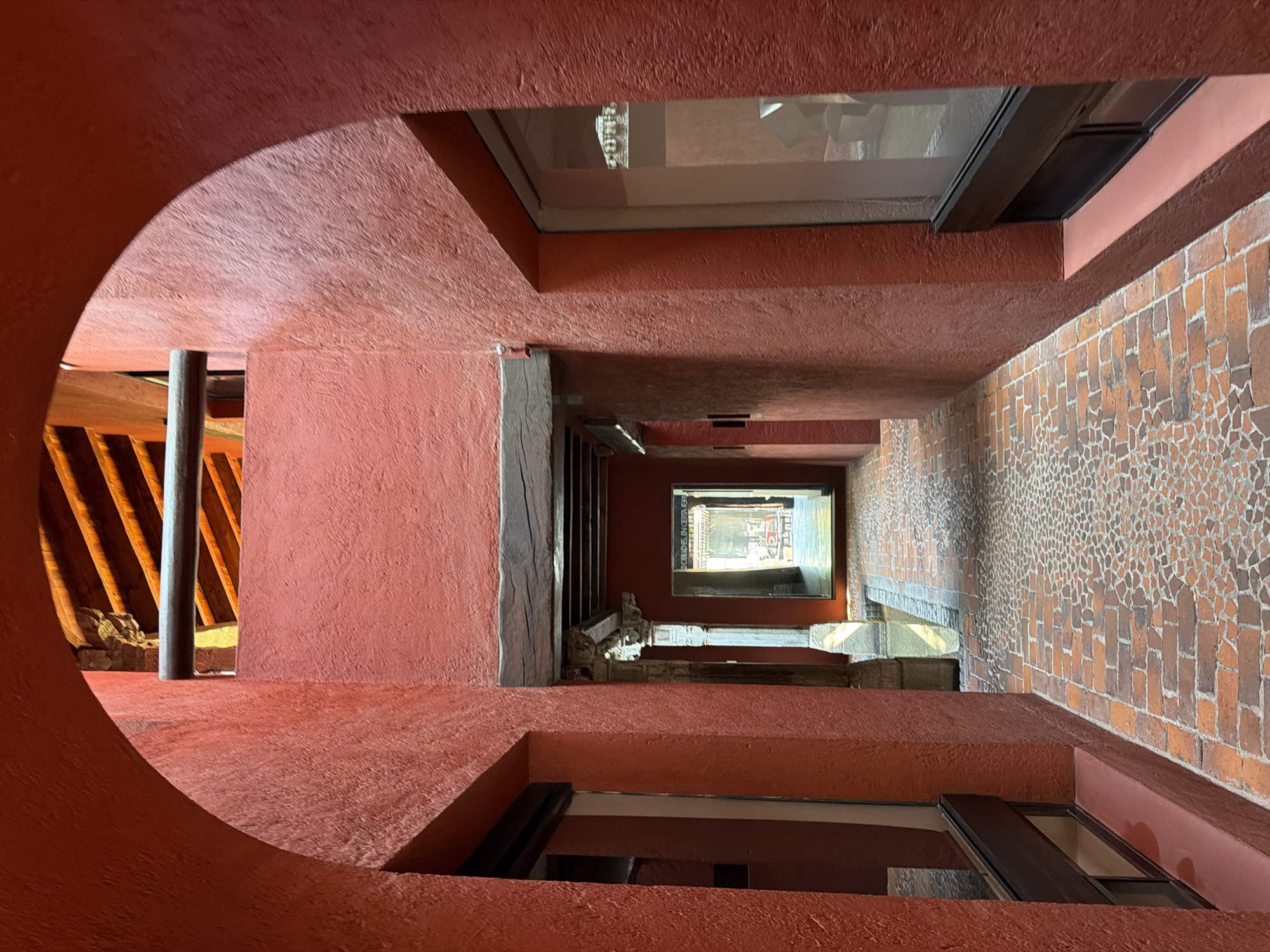 The image captures an interior view of the Museo Amparo in Puebla, Mexico, specifically an architectural detail of the building's structure. The perspective is angled, looking down a corridor with red, textured walls and ceiling. There are several rectangular openings, some containing windows that offer views of the exterior and other parts of the building. The walls feature a mosaic brick pattern. The space has a unique architectural design with arches and angular shapes.
