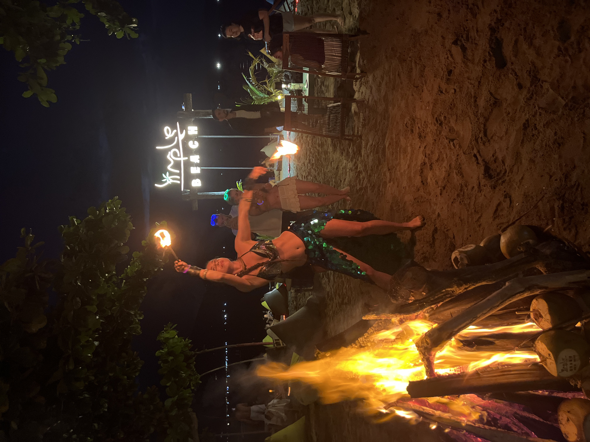The image depicts a nighttime scene on a sandy beach, likely in Đà Nẵng, Vietnam. The main subject is a woman performing a fire-spinning act. She is dressed in a sparkly bikini top and iridescent green shorts, with glowing accessories on her wrists. She is holding a flaming prop in her left hand, and another fire prop is visible in her right hand. She is positioned in front of a large bonfire, which illuminates the sandy ground and surrounding area with a warm, orange glow.
Behind the performer, a neon sign is visible, spelling out "Jungle Beach" in white and blue lights. The sign is mounted on a wooden structure. Several people are visible in the background, some seated at tables and chairs, suggesting a restaurant or bar setting. There are also trees with dark green foliage on the left side of the frame, contributing to the beachside atmosphere. The overall mood is festive and energetic, typical of a beachside entertainment event at night. The sky is dark, indicating it is nighttime, and the presence of flames and minimal lighting suggests no specific weather conditions are evident. The scene is vibrant, with the fire and neon lights creating a dynamic visual contrast against the dark night.