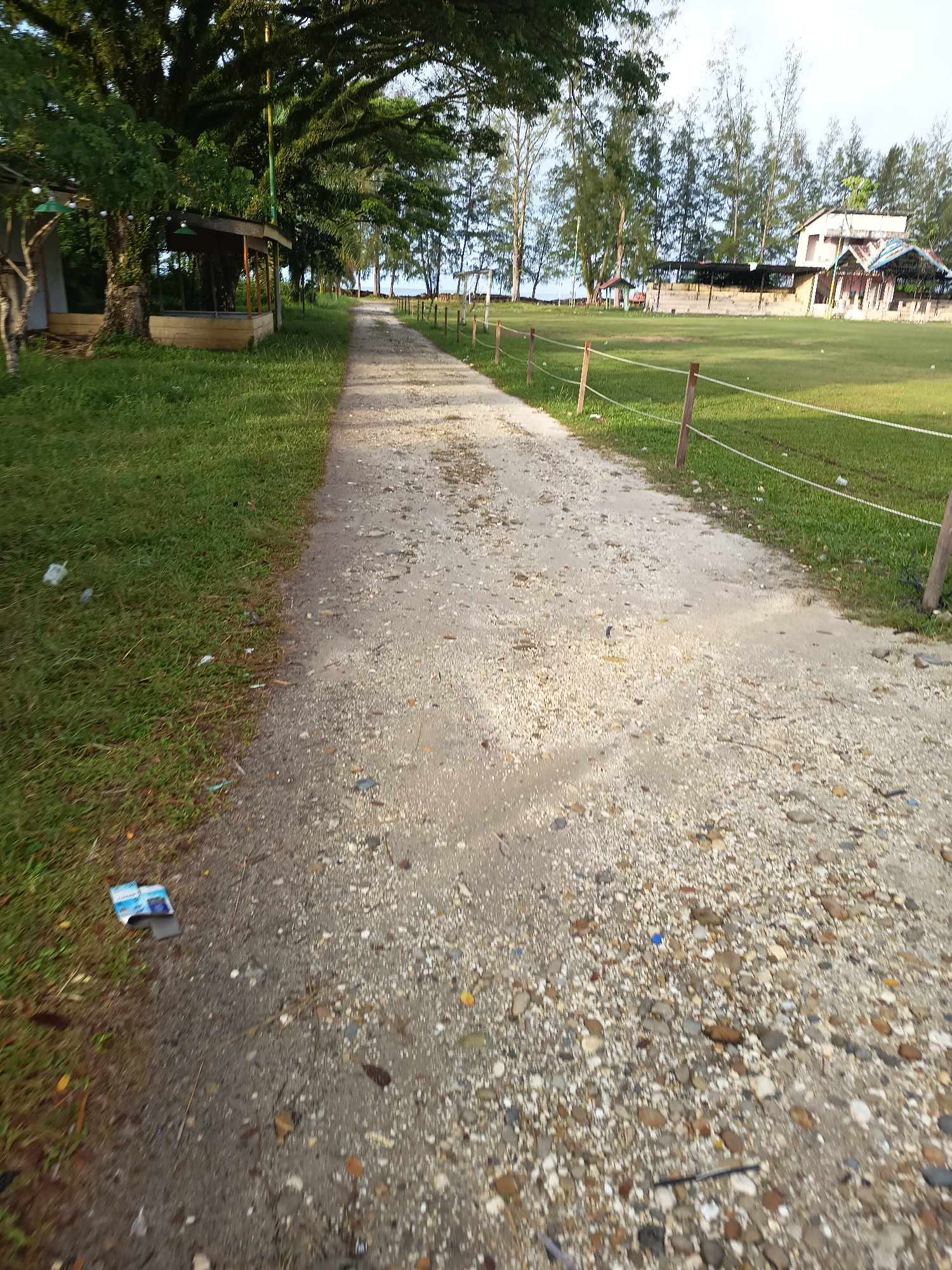 The image depicts a sandy path lined with grass and trees, leading towards buildings in the distance. The path is speckled with small stones and some litter is visible on the grass to the left. In the background, there is a grassy field with a fence, and several structures, including what appears to be a building with a blue roof and another with a white facade. Tall trees with sparse foliage are scattered throughout the scene, and the sky is overcast. The overall impression is one of an outdoor area, possibly a park or recreational space, located in or near the city of Singkil, Indonesia. There are no people visible in the image.