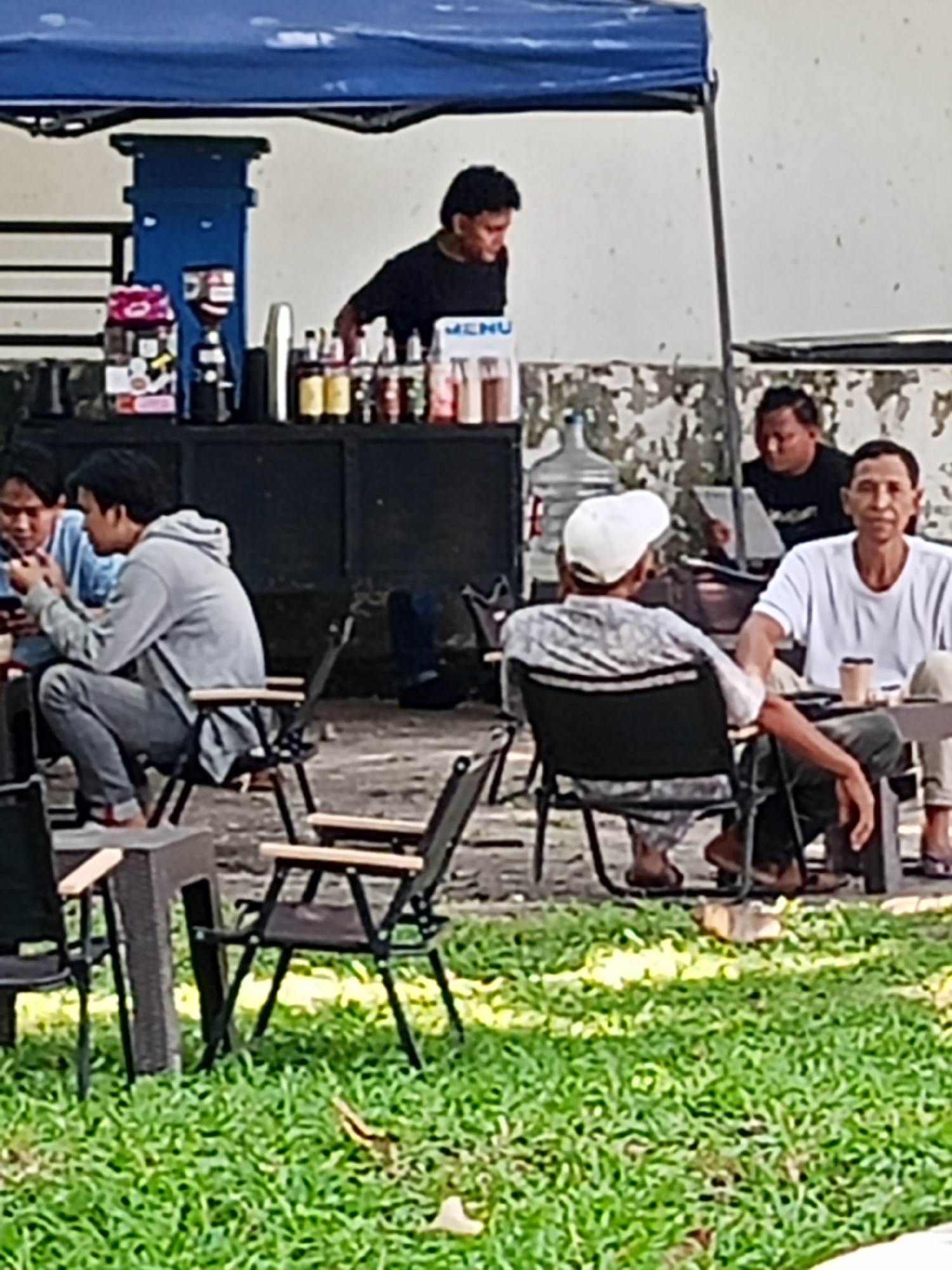 The image depicts an outdoor seating area, likely a cafe or food stall, with several people present. The scene is set against a backdrop of a building with peeling paint, and a blue tarp stretches overhead, providing shade. In the background, a man in a black shirt stands behind a counter, appearing to be serving drinks. On the counter, there are various bottles and containers, including what looks like a coffee grinder.
In the foreground, several individuals are seated on foldable chairs around small tables. One man, wearing a grey hoodie and jeans, is seated and looking at his phone. Another man, wearing a white t-shirt, is seated and looking towards the viewer. A third man, wearing a patterned shirt and a white cap, is also seated, with his back partially to the camera. The ground is covered in green grass, and some dirt paths are visible. The overall impression is of a casual outdoor gathering or a small business operating in an open-air environment. The mention of Lubuk Pakam, Indonesia, suggests a specific geographic context for this scene.