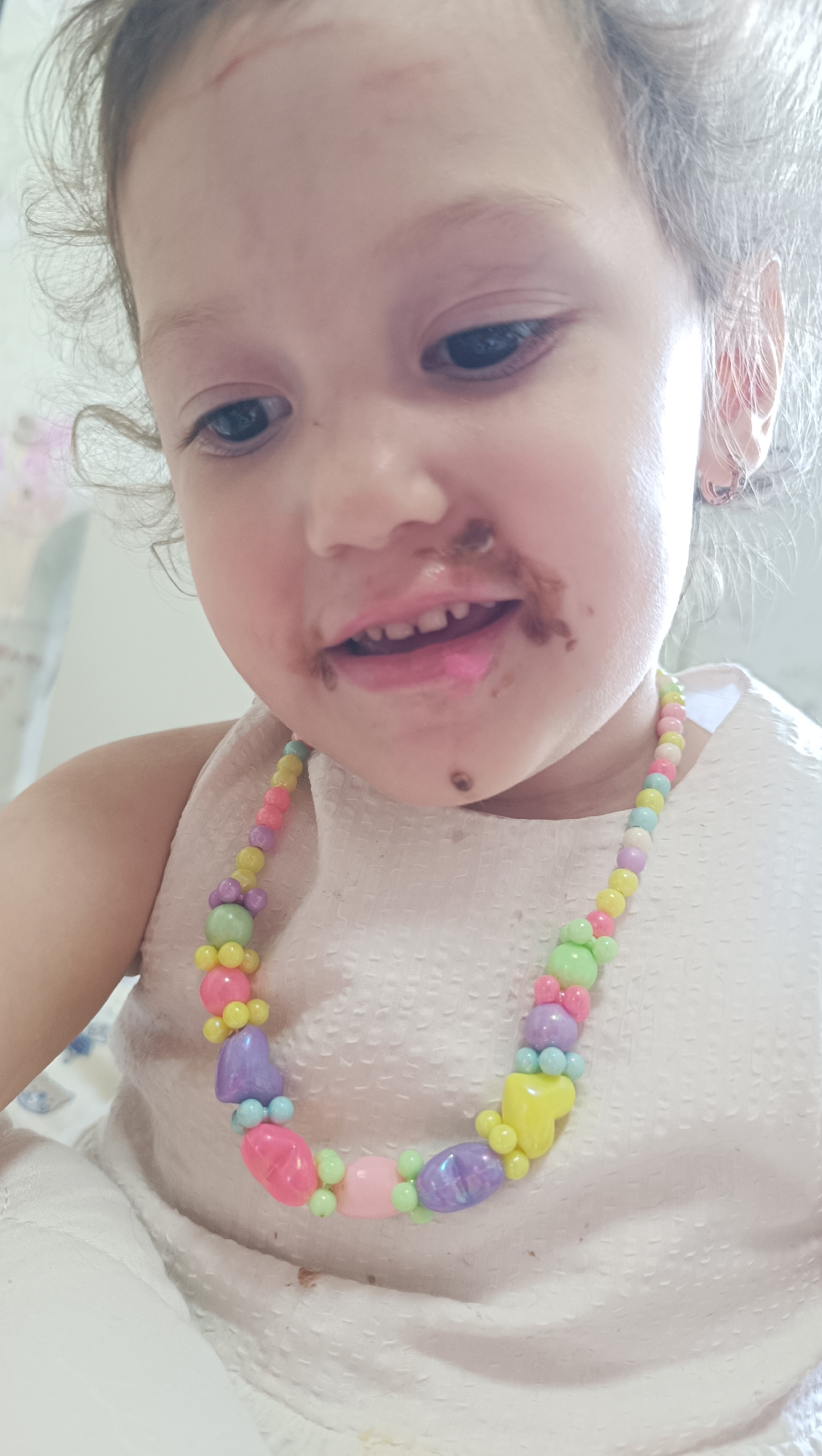 A close-up, low-angle shot shows a young child's face, smudged with chocolate around the mouth. The child wears a white, textured dress and a colorful, beaded necklace. The child's hair is curly and light brown. The background is blurred and appears to be indoors. No specific location in Chaikovsky, Russia, is identifiable from the image.