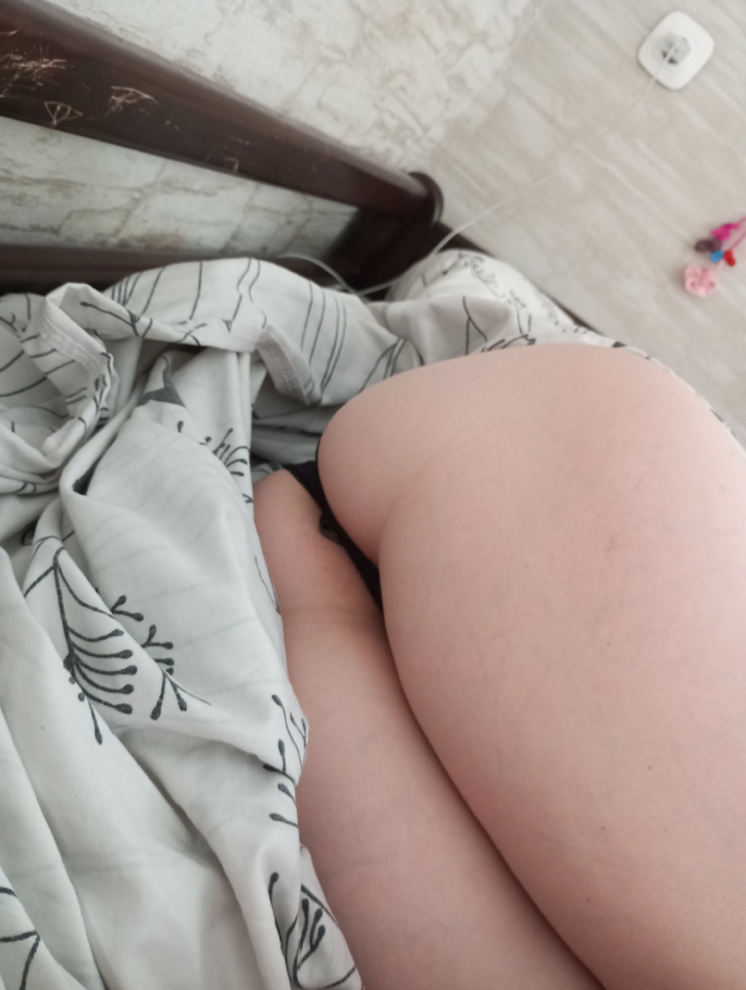 The media file is a close-up, horizontal shot of a person's legs and buttocks in a bed. The person is lying on their side, with their legs bent and their buttocks facing the camera. They are covered with a white duvet that has a black floral pattern. The background shows a tiled wall and floor, with a dark brown headboard of the bed visible at the top left. A white electrical outlet with a cord is on the floor to the right, along with a few colorful small objects, possibly toys or charms. The lighting suggests it is daytime, but the indoor setting makes it difficult to determine the exact time of day or weather conditions. No text is visible in the image. The overall impression is intimate and personal, focusing on a moment of rest or relaxation.