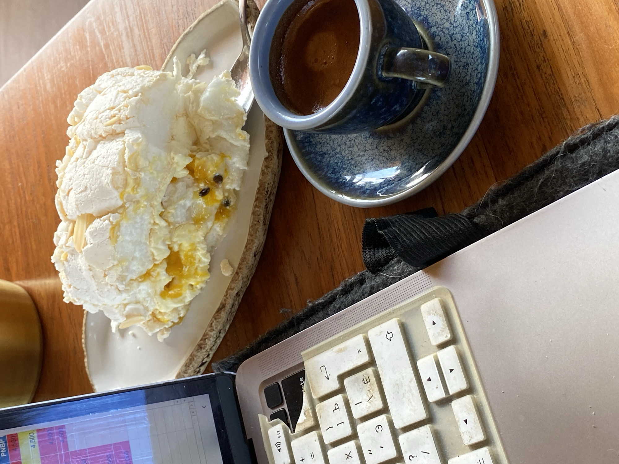 The image shows a close-up view of a table setting with a laptop, a dessert, and a cup of coffee. The laptop is open, displaying a spreadsheet with financial data, suggesting a work or study session. To the right of the laptop is a slice of meringue-based dessert, likely a pavlova, with a filling of passionfruit or a similar yellow fruit pulp and seeds. The dessert is presented on a rustic-looking ceramic plate. Adjacent to the dessert is a small, dark blue ceramic cup filled with dark liquid, presumably coffee, resting on a matching saucer. The table is made of wood with a visible grain. The scene is illuminated by natural light, casting soft shadows. The setting appears to be indoors, possibly a cafe or a personal workspace, in Kuta Selatan, Indonesia. No people are visible in the image. The overall atmosphere is one of focused work accompanied by a sweet treat and coffee.