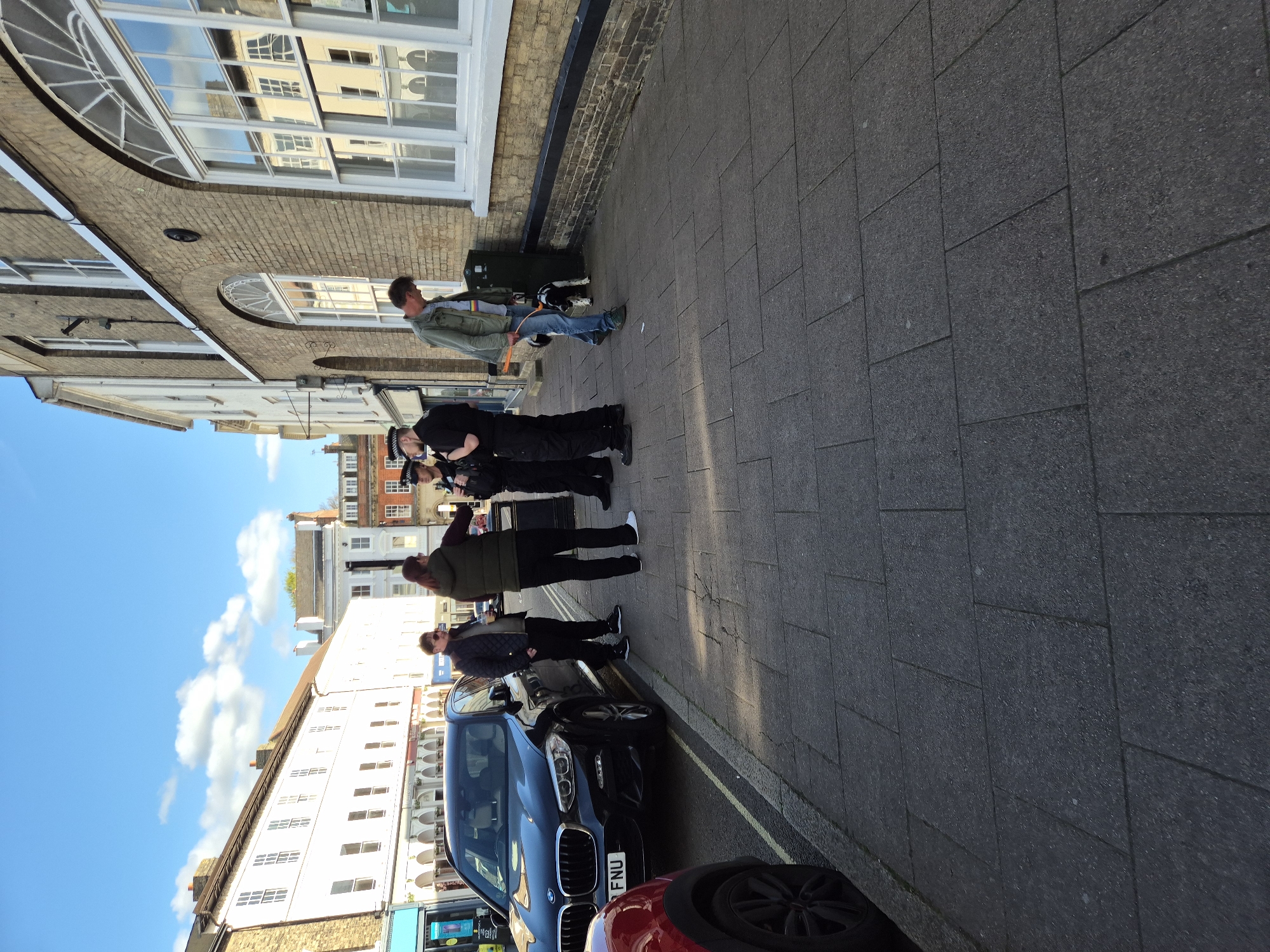 The image shows a street scene in West Suffolk, United Kingdom, with several people and vehicles present.
Four people stand on the paved sidewalk. The first person from the left is wearing a plaid jacket and black pants. Next to this person stands someone wearing a green jacket and black pants. Next to the second person, there are two police officers in uniform, one of whom is taking a photo of someone. A man wearing a green jacket and jeans stands with a dog next to the building on the sidewalk.
Several cars are parked along the curb, including a blue BMW with license plate FNU. Buildings with multiple windows are visible on the left, with a blue sky and clouds in the background. A brick building with large windows is on the right. The sidewalk is made of gray rectangular stones.
