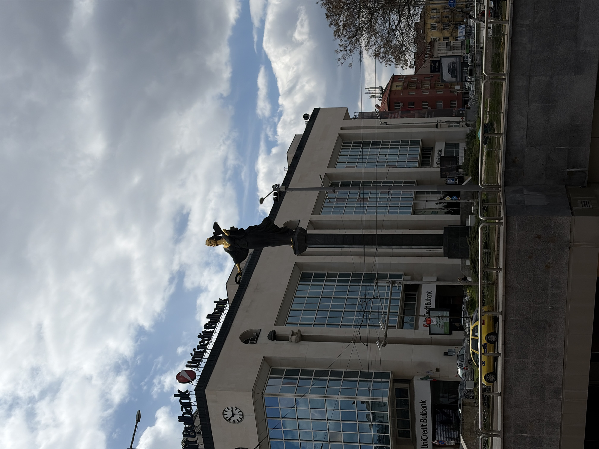 The image shows the Statue of Sveta Sofia in Sofia, Bulgaria. The bronze statue of a woman stands atop a tall pillar. The statue and pillar are positioned in front of a multi-story building with a clock, signage for UniCredit Bulbank, and numerous windows that reflect the cloudy sky. A street with parked cars is visible in front of the building, and a barrier of metal rails separates the street from the building's facade. In the background, there are additional buildings and trees. The sky is partly cloudy.