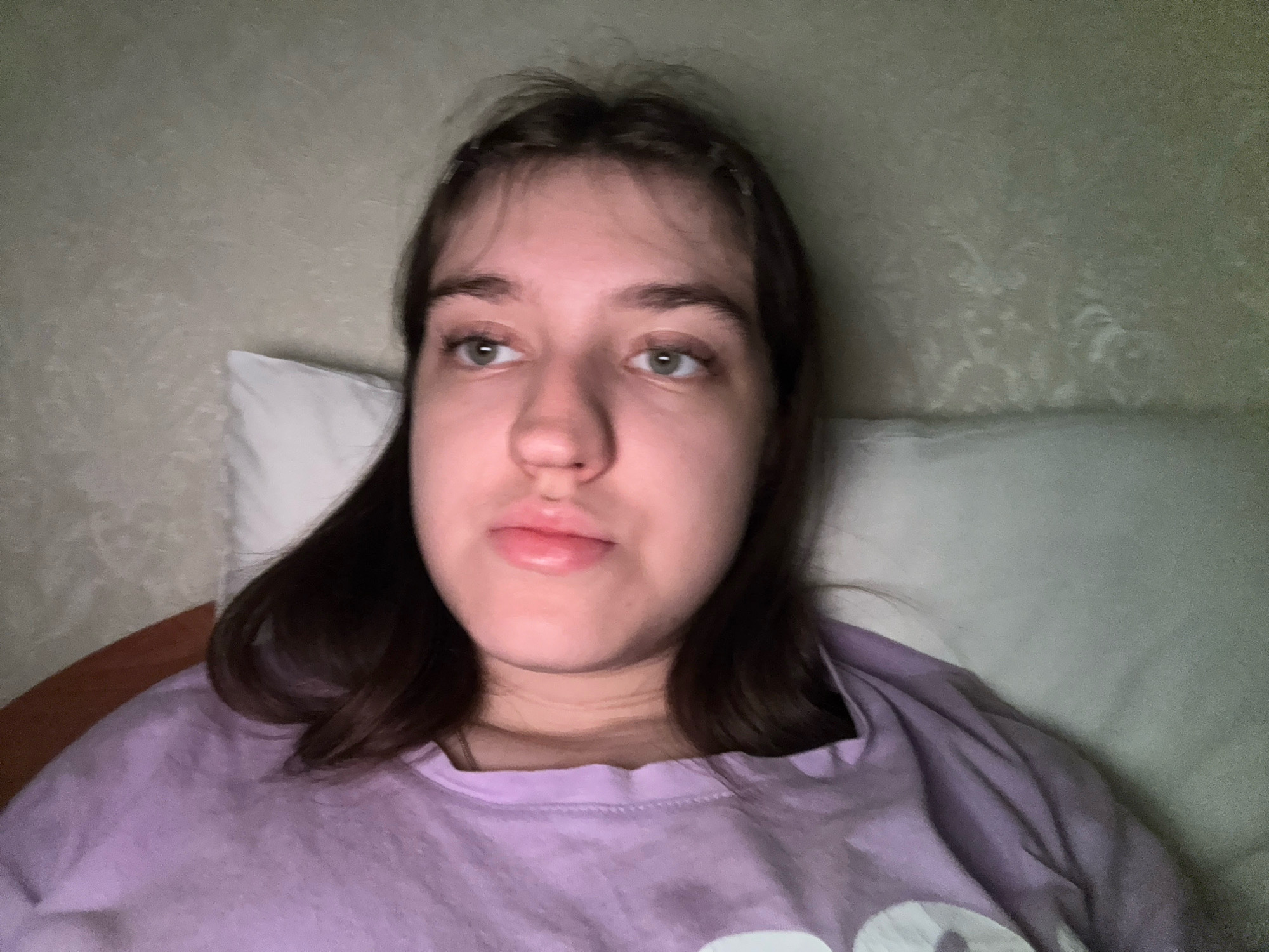 A young person with brown hair and light skin is lying down, wearing a purple t-shirt. They are looking to the side with a neutral expression. The background consists of a textured wall and a white pillow. The lighting is dim and appears to be artificial, casting soft shadows. The scene suggests an indoor setting, possibly a bedroom, in Zavolzhye, Russia.