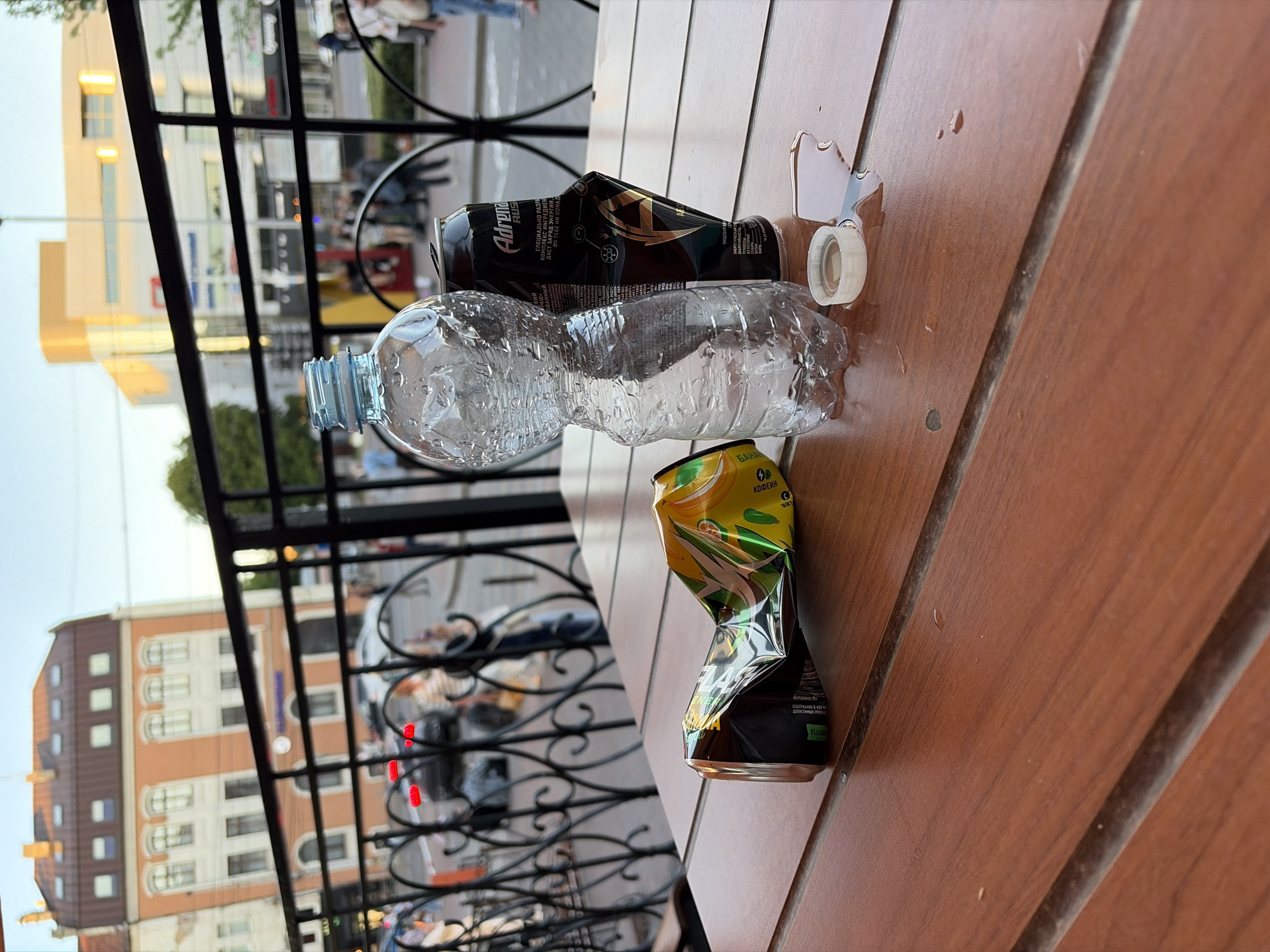 In this medium shot, a wooden table in what appears to be an outdoor seating area of a cafe or restaurant is the primary focus. The table's surface is a warm, reddish-brown with visible wood grain and horizontal slats. On the table, two crumpled drink containers are present: a crushed yellow and green can to the left, and a partially crushed black can to the right, bearing the "Adrenaline Rush" logo and Russian text. Beside the black can stands a clear plastic water bottle, partially filled with water, which has condensed droplets on its surface. The bottle's cap lies to the right of the bottle, adjacent to a small puddle of liquid on the table.
In the background, a black metal railing with decorative scrollwork separates the seating area from a street scene. Beyond the railing, several blurred figures can be seen, including people walking and what appears to be a parked car. The environment suggests an urban setting, with buildings visible in the distance. The closest building is a multi-story structure with orange and white facade and numerous windows. Further back, a modern building with a cream-colored facade and large windows is visible, catching the light. The overall lighting suggests it is daytime, possibly late afternoon or early evening, given the warm tones and shadows. The image is slightly tilted, giving a dynamic perspective to the scene.