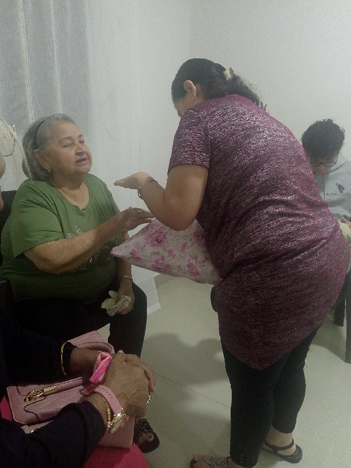 An older woman in a green shirt sits facing another woman standing and holding a floral pillow. The seated woman gestures with her hands. A third person is partly visible seated beside the older woman. The standing woman wears a sparkly maroon top and black pants. A young person with short dark hair and glasses sits in the background, wearing a light gray sweatshirt. The scene takes place indoors, with a curtain visible on the left side of the image.