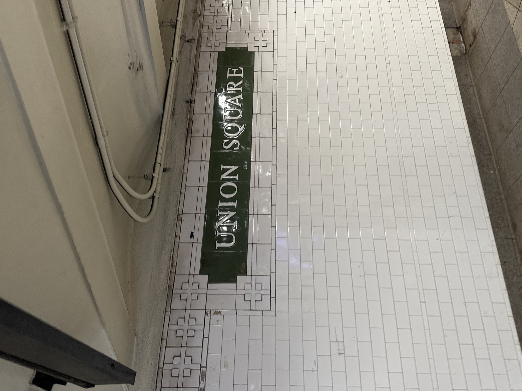 The image depicts a section of an underground subway station, likely in New York City given the context and visible text. The photograph is oriented vertically, showing what appears to be a tiled wall on the right and a tiled platform or floor on the left, with an additional beige wall further left.
A prominent feature is a dark green tiled sign embedded into the white rectangular subway tiles, displaying the station name "UNION SQUARE" in large, white, serif capital letters. The white tiles around the green sign feature some decorative patterns composed of smaller squares. The main white tiled surface shows visible grout lines and some minor scuffs or dirt, indicating wear.
To the left of the tiled area, a beige-colored wall is visible, with several white pipes or conduits running along its surface. These pipes appear to be part of the station's infrastructure. On the far right edge of the image, a darker, possibly granite or concrete, textured surface suggests the edge of the platform bordering the tracks.
No people are present, and no specific activity is taking place. The scene is static, capturing an architectural detail of the subway environment. The lighting is consistent with an indoor, artificially lit setting. The overall impression is one of a functional, well-used public transport space.