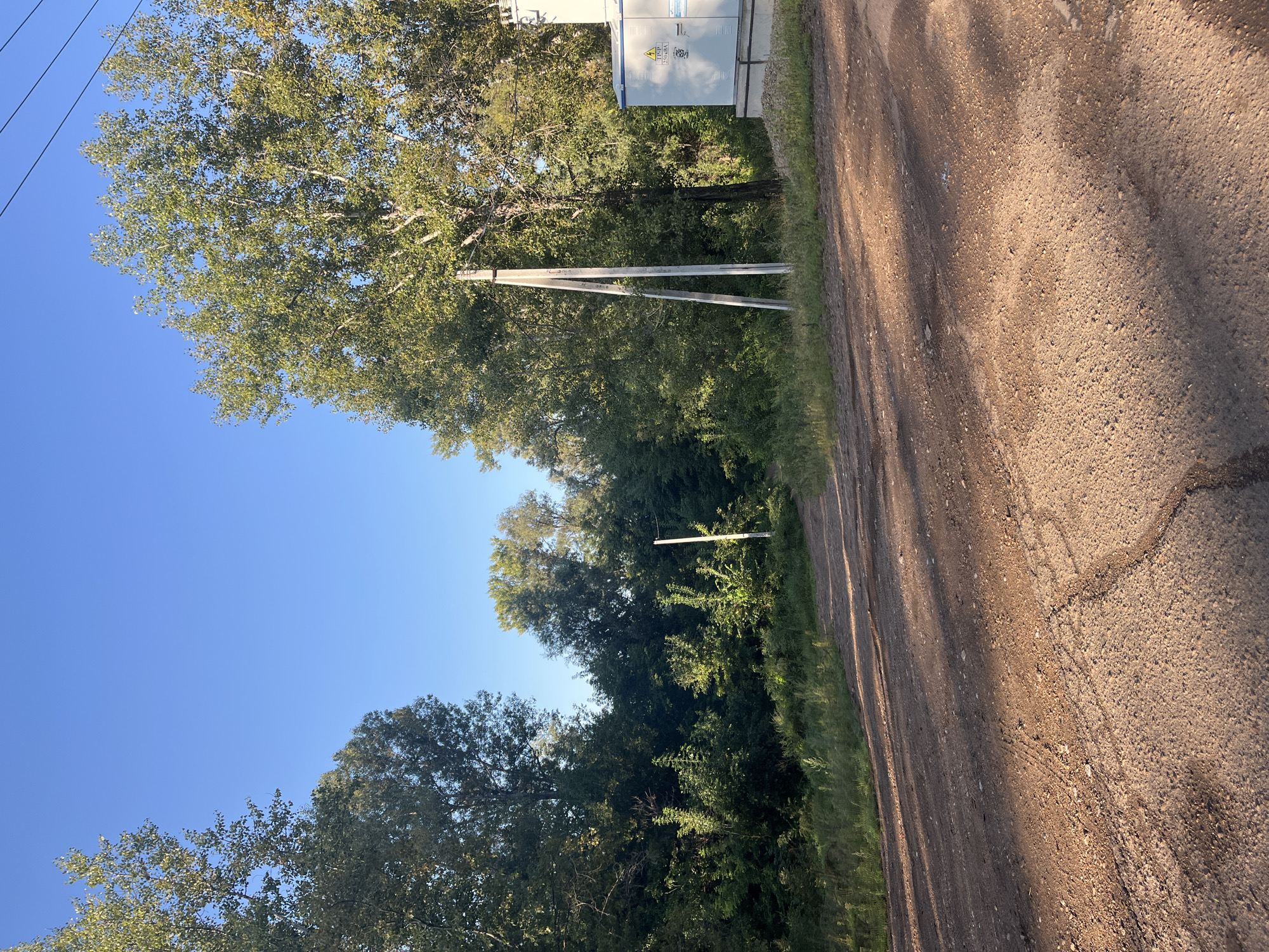 The image depicts a dirt road winding through a rural landscape with lush green trees on either side. The sky above is a clear, bright blue. In the foreground, the road is textured with gravel and shows signs of wear, with tire tracks visible. Sunlight casts long shadows across the road from the trees. On the right side of the image, a light-colored utility box or substation is visible, and two tall, light-colored concrete poles stand nearby, likely supporting electrical lines which are also visible against the sky. The overall impression is of a natural, undeveloped area, possibly on the outskirts of a town or city. The mention of Blagoveshchensk, Russia, suggests this location is in that specific geographical area, which is known for its natural landscapes and proximity to the Amur River. However, there are no specific landmarks in the image that definitively confirm this location. There are no people or animals present in the scene.