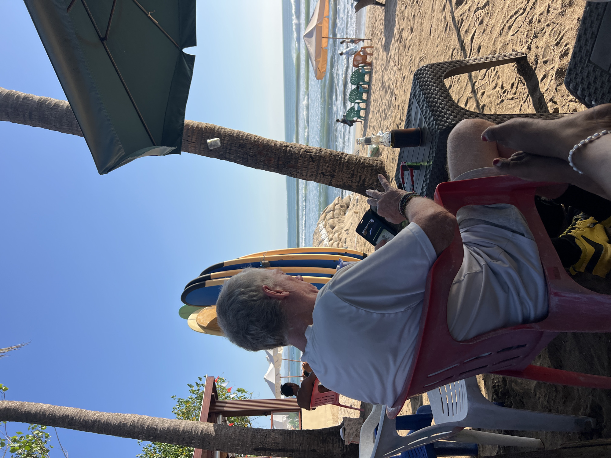 The image captures a beach scene in Kuta, Indonesia, under a clear blue sky. In the foreground, a man with grey hair is seated in a red plastic chair, facing away from the camera and looking at a tablet. He is wearing a white t-shirt and light-colored shorts. A dark-colored beverage bottle and the tablet are on a woven plastic table in front of him. To his left, a white plastic chair is visible.
Behind the man, several surfboards are propped up against a structure that appears to be part of a beachside establishment, possibly a cafe or rental shop. A palm tree trunk is prominent on the left side of the frame, with lush green foliage visible above. Another larger palm tree trunk is positioned to the right of the surfboards, with a dark green umbrella extending from its upper portion.
Further into the scene, the sandy beach stretches towards the ocean, where gentle waves are breaking. Several beach umbrellas and chairs are scattered on the sand. Two people are visible near the water's edge, and another person is seated in a red chair under an umbrella. The ocean stretches to the horizon under the bright sky. The overall atmosphere suggests a relaxed beach environment.