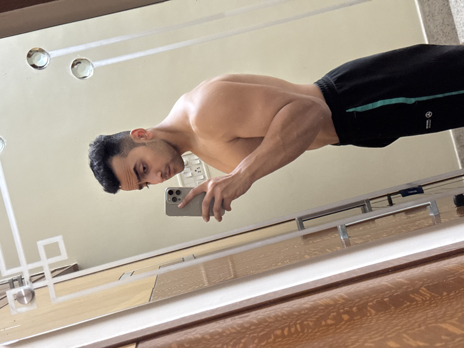 A muscular man takes a mirror selfie. He has dark hair and is shirtless, wearing black athletic shorts with green and white accents. He holds a silver-toned smartphone, partially obscuring his face. The reflection showcases a cream-colored wall with decorative mirror details. The foreground features a wooden surface with visible grain, likely a vanity or dresser.