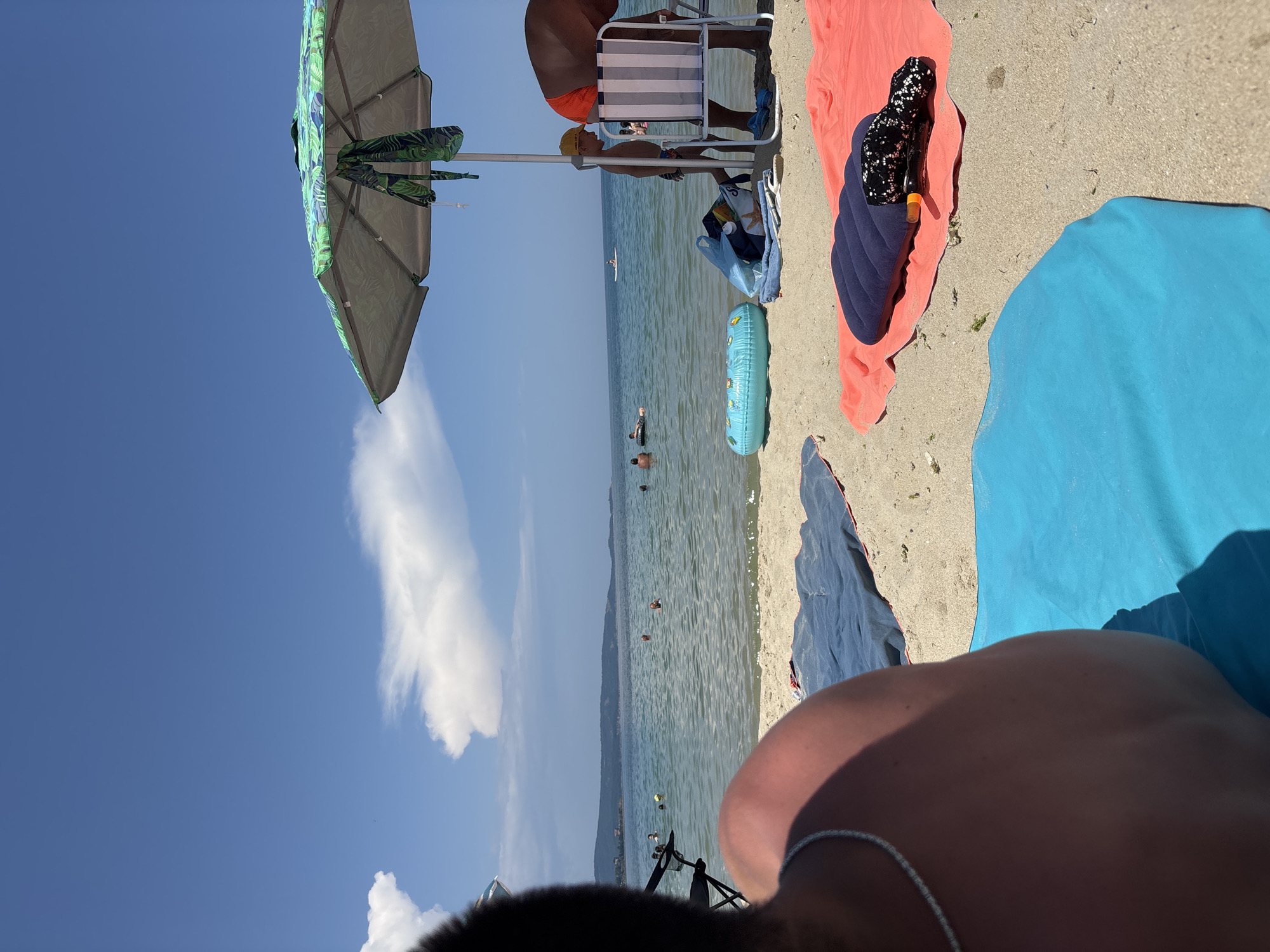 The image depicts a sunny day at a beach in Kiten, Bulgaria. The foreground shows several beach towels laid out on the sand: a large blue towel, a gray towel, a coral-colored towel, and another blue towel. On the coral towel, there is a deflated navy blue inflatable cushion and a black and white patterned cloth. Behind the towels, the calm sea stretches towards a distant, hazy coastline with hills. Several people are visible in the water, some swimming and others using inflatable rings. A large, green and white patterned beach umbrella provides shade. Underneath the umbrella, a child wearing a yellow cap stands next to a striped beach chair. To the right, the back of a person wearing orange swimming shorts is visible. The sky is a clear blue with a few white clouds.
