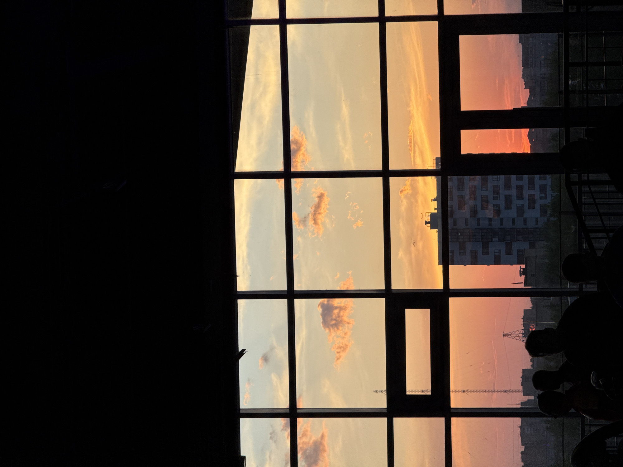 The image depicts a view of a sunset seen through a window, possibly in an urban setting. The window is divided into rectangular panes. Beyond the window, the sky displays a gradient of colors, from yellow to orange, suggesting a sunset or sunrise. Silhouettes of buildings and a transmission tower are visible in the distance against the colored sky. A dark silhouette appears on the left side of the image, and a group of silhouetted heads and shoulders are present on the right side of the window. The image was taken in the city of Krasnoyarsk, Russia.