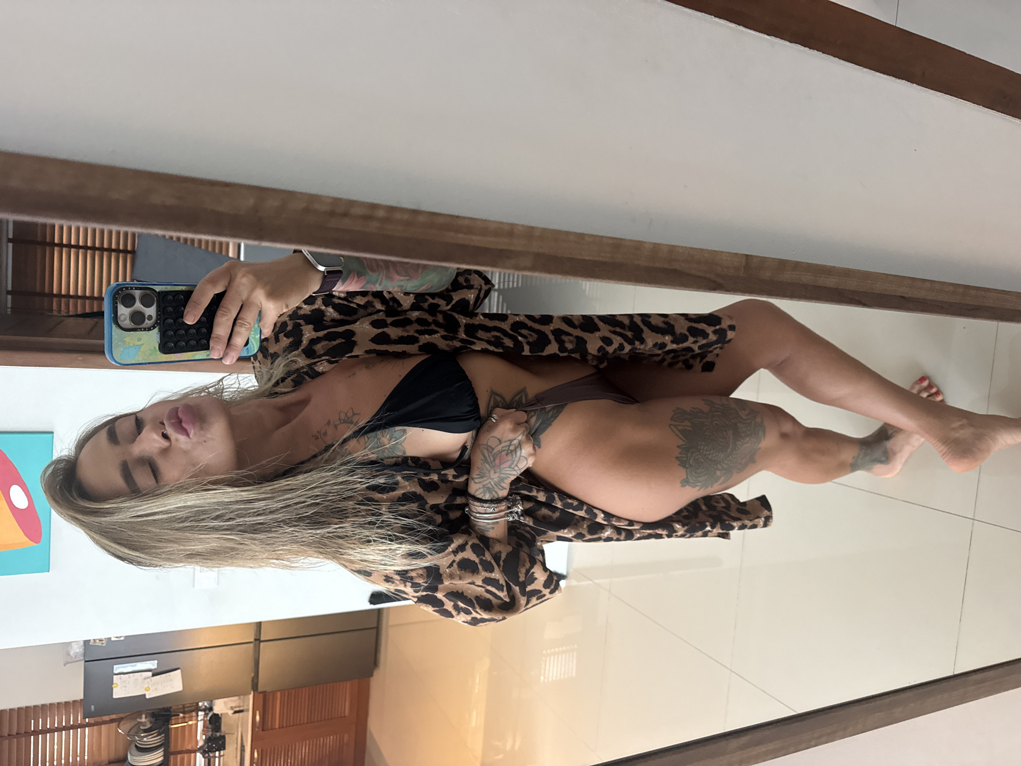 This media file captures a woman taking a mirror selfie. She is wearing a leopard print robe over a black bikini. The woman has blonde hair and numerous tattoos on her arms and legs. She is posing with her lips pursed. The background shows a modern interior with tiled flooring and wooden accents. A colorful piece of art is visible on the wall to the left. The lighting suggests it is daytime. The setting appears to be a residential or hotel room in Thep Krasatti, Thailand. The woman's pose and facial expression convey a playful or flirtatious mood.