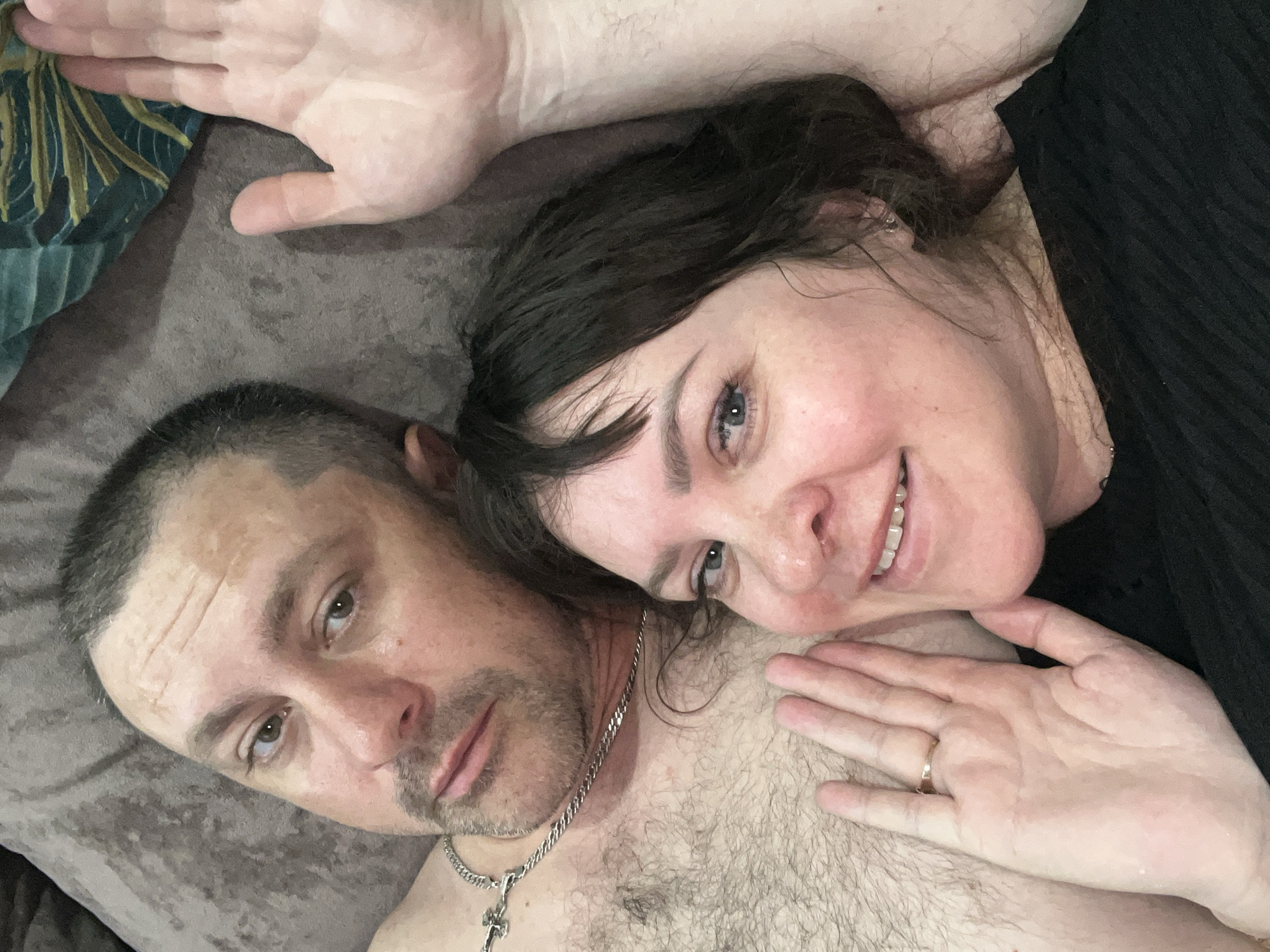 A close-up, angled selfie depicts a Caucasian couple lying on what appears to be a brown upholstered surface. The man has short, graying hair, light-colored eyes, stubble, and a silver chain with a cross pendant. He is shirtless, revealing chest hair. The woman has dark hair, fair skin, and blue eyes, and she is smiling slightly. Her hand rests on the man's chest, and she is wearing a ring on her ring finger. An additional hand is partially visible in the upper left corner of the image. The exact location in the city of Noviy, Russia, cannot be determined from the image.