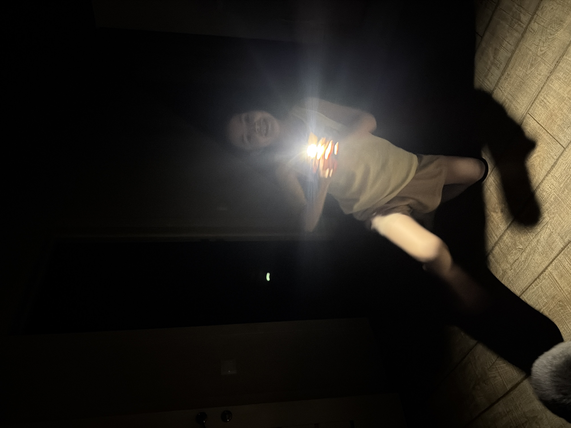 This is a portrait-oriented, low-light photograph with a strong flashlight beam illuminating the scene from the center. The primary subject is a person, likely a young girl, who is lying down and holding a small, bright object, possibly a flashlight or a light-up toy, directly in front of her face. The light from this object is intensely bright, creating a lens flare and obscuring some details of the girl's face, though her smiling expression is discernible. She is wearing a light-colored tank top and shorts.
The background is predominantly dark, suggesting nighttime or a dimly lit indoor space. To the right of the girl, the floor is visible and appears to be made of light-colored wooden planks, laid diagonally. A long, dark shadow stretches across the floor from the girl, cast by the flashlight's beam. In the far background, a faint, small, crescent-shaped light source is visible in the darkness, its nature unclear. The overall mood is playful and adventurous, perhaps suggesting a game of flashlight tag or storytelling in the dark. No text is visible in the image. The location context is Шмидтово, Russia.