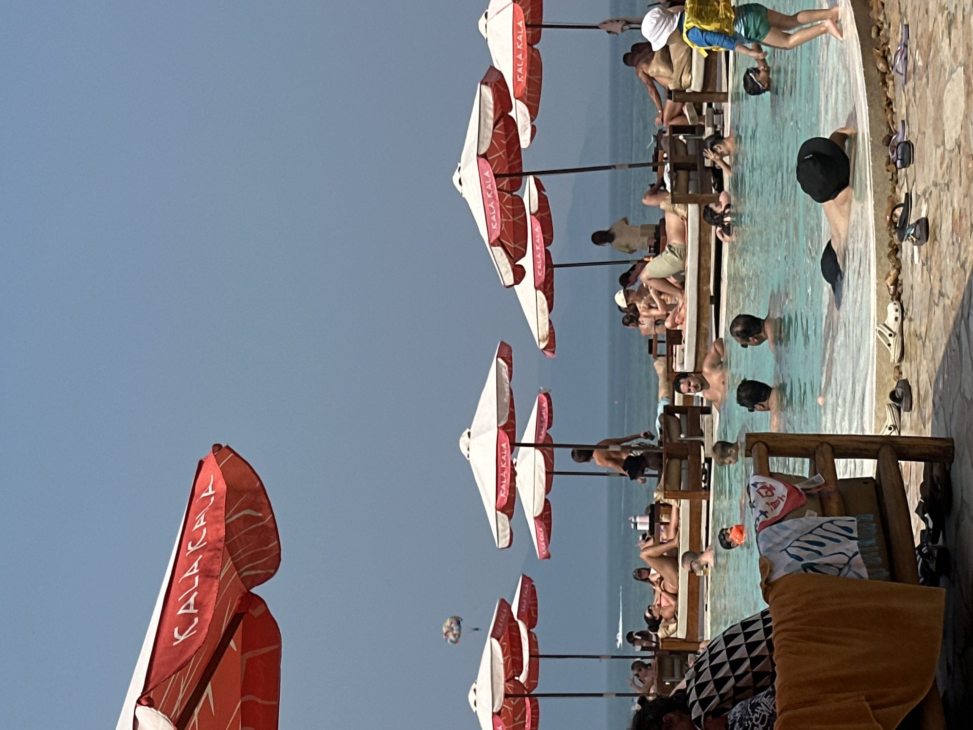 The image depicts a beach scene, possibly near Đà Nẵng, Vietnam. The scene features a swimming pool or beach club setting with numerous people engaging in leisure activities such as swimming, sunbathing, and lounging on chairs. Red and white branded umbrellas ("KALA KALA") provide shade. In the distance, the sea is visible, and there is a parasailing activity occurring.