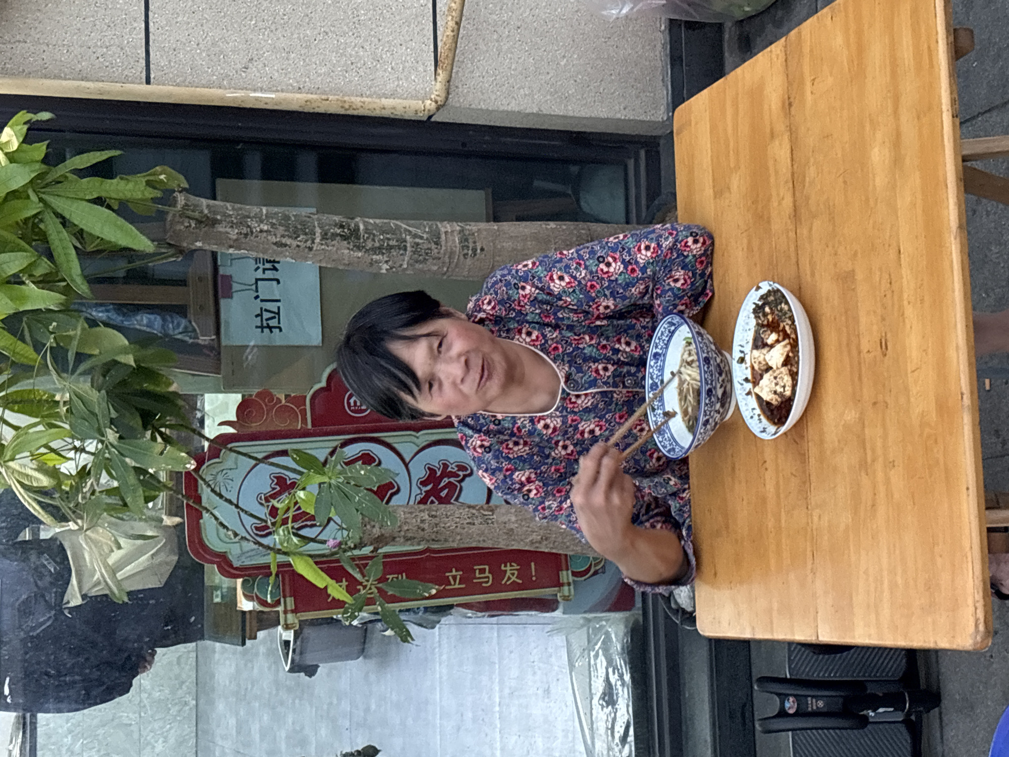 A person with short black hair and wearing a floral patterned shirt is seated at a wooden table outside, holding chopsticks. Two bowls are on the table, one appearing to contain noodles and the other a sauced dish. In the background, a sign with Chinese characters and the phrase "立马发!" is visible, suggestive of Jiangjin, China. A potted plant stands beside the table.
