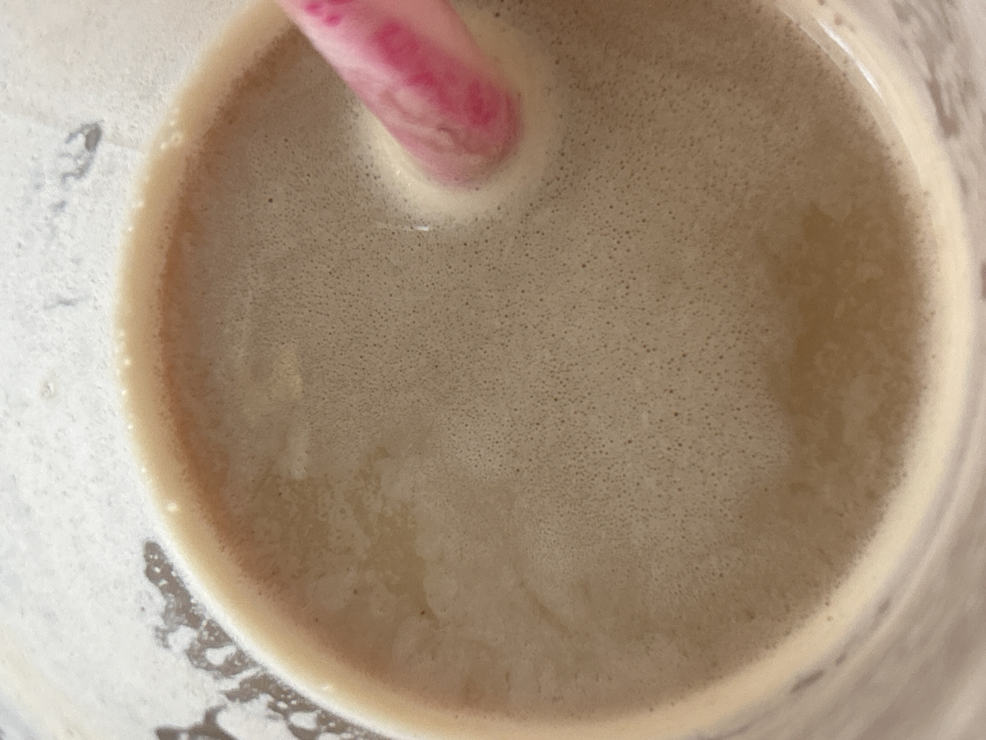 A close-up, top-down view shows the foamy surface of a light brown liquid in a clear container, likely a beverage. A pink straw is partially submerged on the right side, creating a subtle disturbance in the foam. The liquid has a textured appearance due to many small bubbles. The background is indistinct and light. The scene is a close-up of a drink and does not provide any information about a specific location in Plovdiv, Bulgaria, or any other environmental details.
