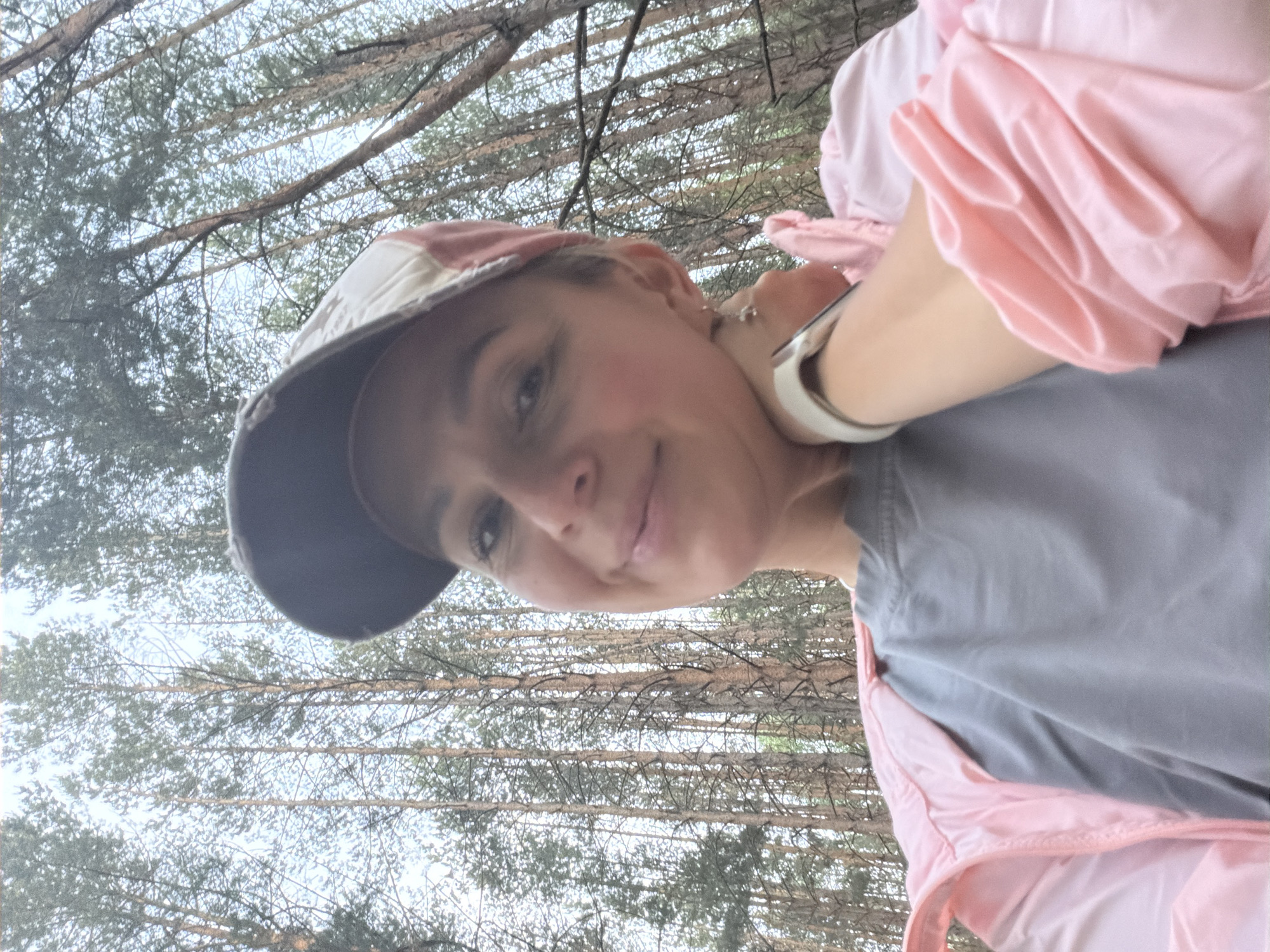 A woman wearing a baseball cap, gray shirt, and pink jacket is taking a selfie in a wooded area. The location is Chaikovsky, Russia. Bare tree branches are visible in the background.