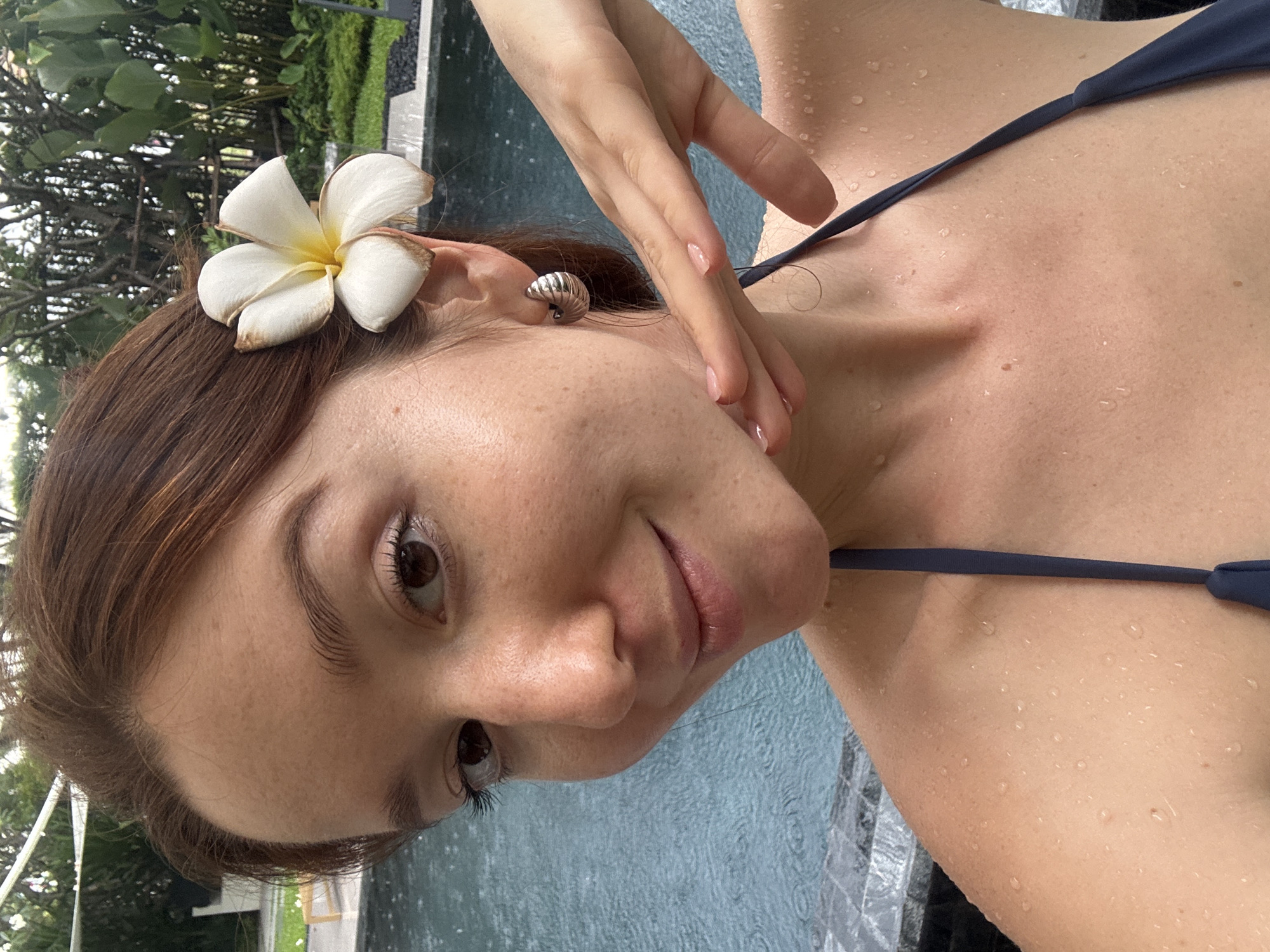 A woman with brown hair adorned with a white flower is posing for a selfie near a pool in Choeng Thale, Thailand. She is wearing a navy blue bikini top and has water droplets on her skin. The environment includes lush greenery in the background and clear pool water.