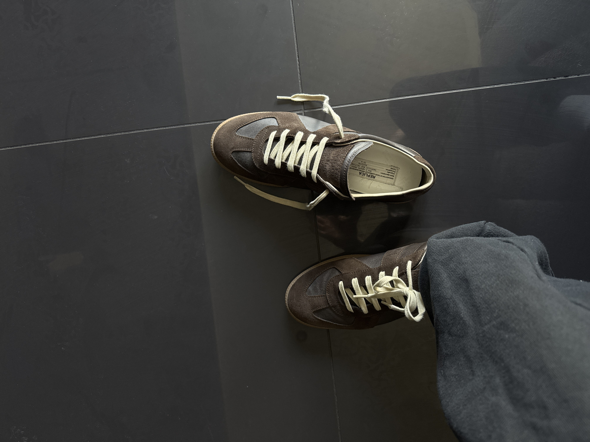 The image shows a pair of brown Maison Margiela "Replica" sneakers on a dark gray tiled floor. One shoe is worn by a person wearing dark gray pants. The setting appears to be indoors. The location is not discernible from the visual information in the image.