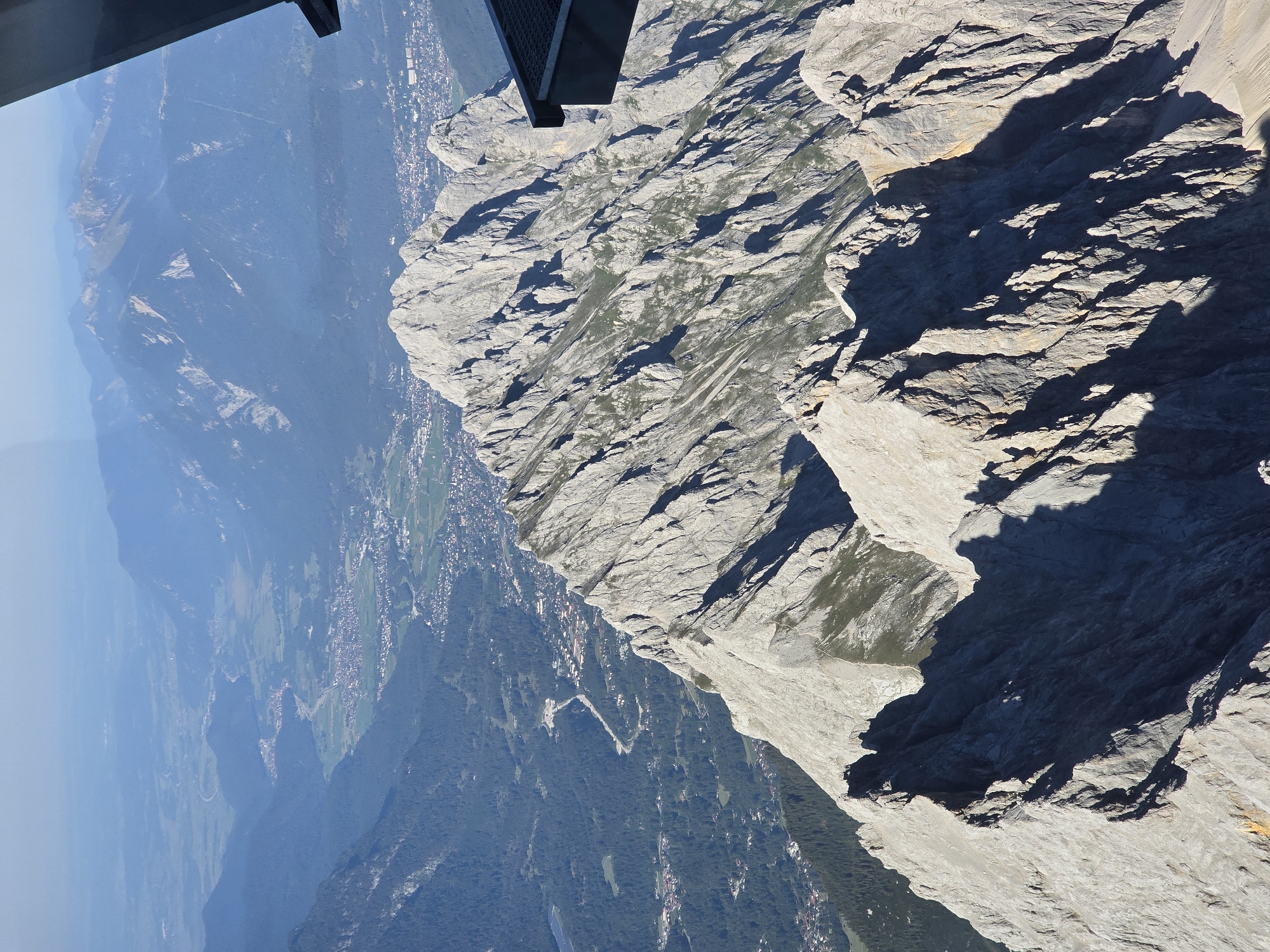 The image is a high-angle aerial view of a mountainous landscape with a town in the valley below. The foreground features rugged, rocky mountain ridges with steep slopes and deep shadows. In the mid-ground, a town with buildings and roads is nestled in a green valley. The background consists of more mountain ranges, stretching into the hazy distance under a clear blue sky. A dark, geometric structure, possibly part of a viewing platform or building, is visible in the upper right corner, suggesting the vantage point is from a man-made structure. The image captures a panoramic vista of the Bavarian Alps, with the town likely being Garmisch-Partenkirchen, Germany, given the dramatic mountain scenery. There are no people or animals visible in the frame.