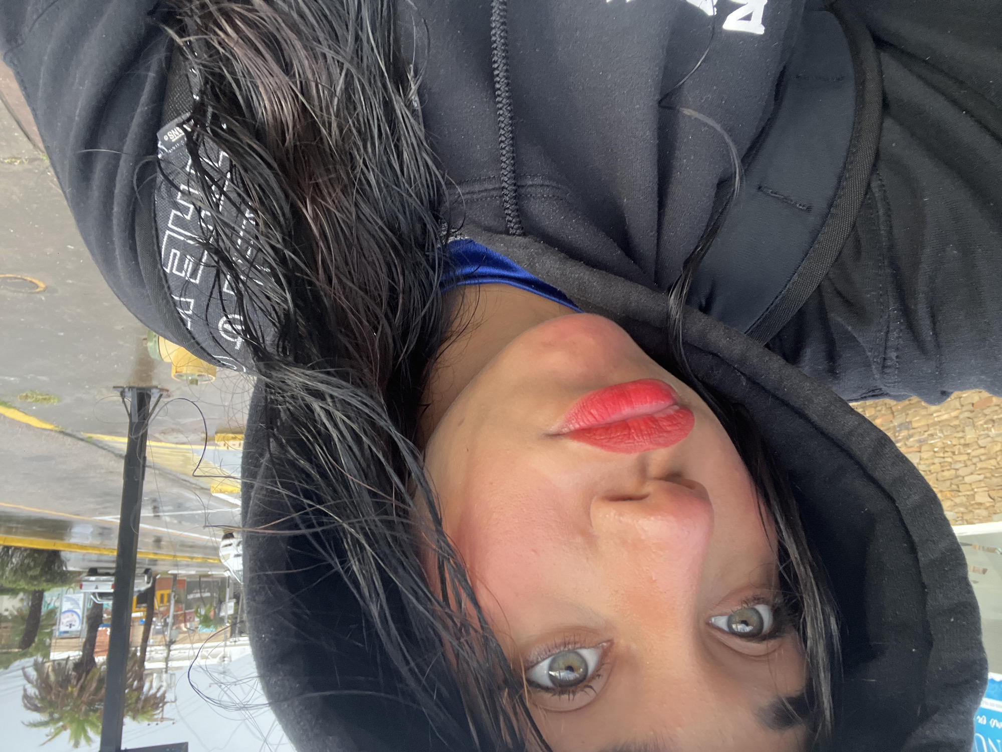 A selfie-style, close-up, upside-down photograph features a woman with wet, dark hair wearing a black hooded sweatshirt. A blue undershirt is visible at the collar. She has fair skin, red lipstick, and light-colored eyes.
The background, also upside-down, suggests an urban environment. Visible elements include a street with yellow lines, a light post, a palm tree, signage, and a vehicle, consistent with Pampatar, Venezuela. The presence of wet hair and wet surfaces suggests a recent or ongoing rain event.