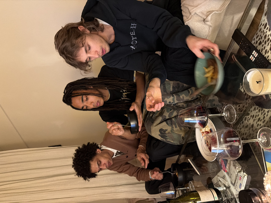 The image depicts three young men engaged in a casual meal within an indoor setting, likely a private residence.
The leftmost individual is a male with dark, tightly curled hair, wearing a brown cardigan over a white collared shirt and a dark tie. He sits with a small black bowl in his left hand and a fork in his right, looking down at his food.
The middle individual is a male with long, dark dreadlocks, dressed in a dark t-shirt and camouflage-patterned trousers. He holds a small black bowl with both hands and a fork, also looking down at his meal.
The rightmost individual is a male with medium-length, light brown wavy hair, wearing a dark hooded sweatshirt with "ACTE H" or similar text on the front, and dark trousers. He holds a teal-colored bowl containing food, appearing to be fries, in his left hand, observing its contents.
All three men are seated around a dark, reflective coffee table, which rests on a light-colored rug with a dark animal print pattern. On the table, multiple clear stemmed wine glasses, mostly filled with red wine, are present. A green glass wine bottle, partially filled, stands uncorked. A white rectangular plate with remnants of food is visible. Other items include a white package or box labeled "Apprazola 20 mg," a burnt white pillar candle in a glass holder, and a black remote control.
The background consists of light-colored walls and beige drapes, suggesting a living room or dining area. The lighting is warm and ambient, without visible natural light sources.
Regarding location, the image provides no specific external or internal landmarks, signage, or architectural cues to definitively verify its placement within Paris, France. The scene is consistent with a private residential interior.