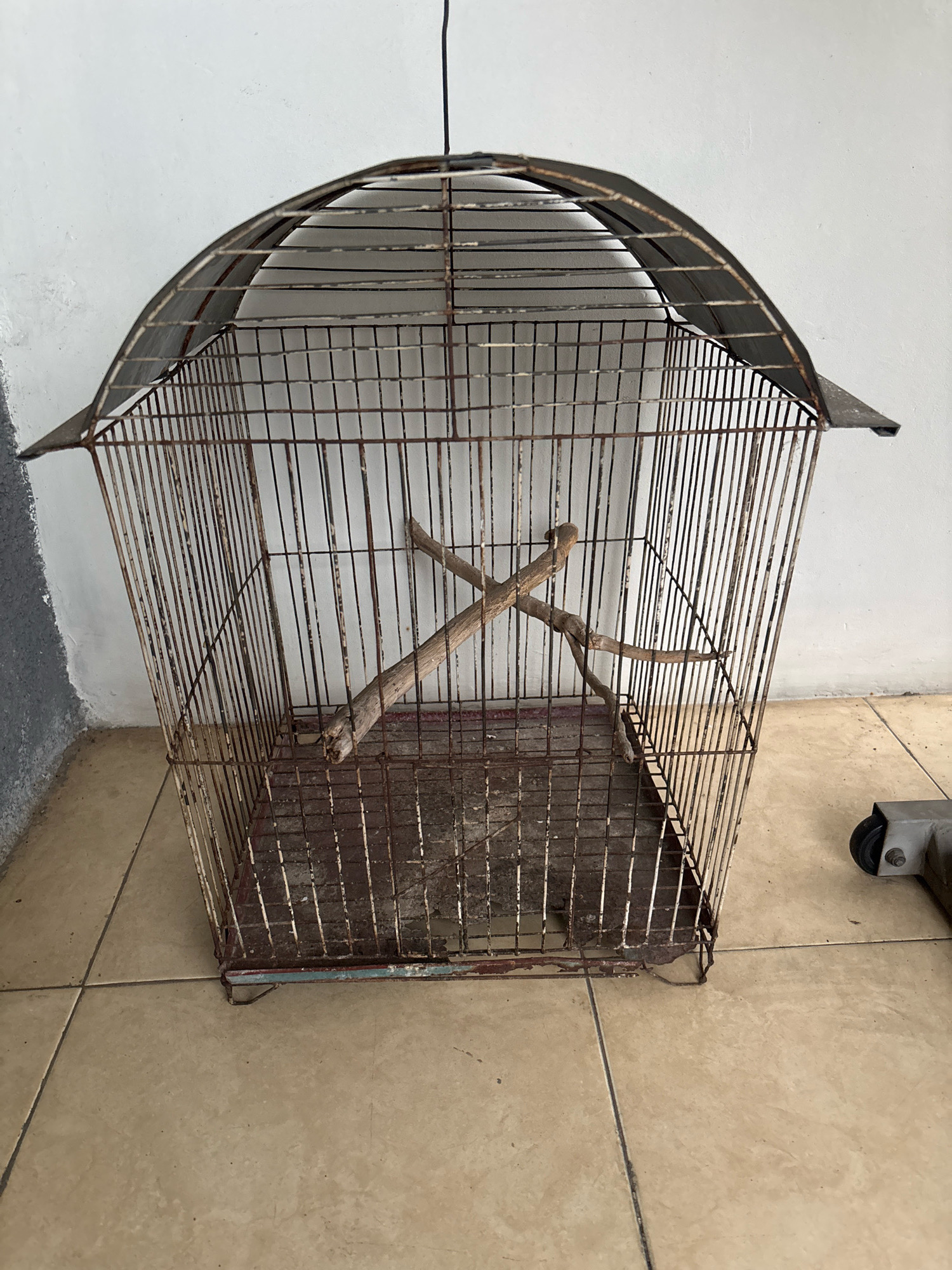 The image displays an empty, aged, and rusty birdcage with two wooden perches inside. The cage has a domed top and a flat base, and it is constructed from thin metal bars. The cage rests on a tiled floor, and a white wall serves as the background. A portion of a metal object with a wheel is visible in the lower right corner of the image. The scene appears to be indoors, possibly in a storage area or a less-maintained part of a building. The lighting is even, suggesting an overcast day or artificial lighting. There are no people or animals present in the image. The setting is likely residential or a small commercial space in Bekasi, Indonesia, given the context.