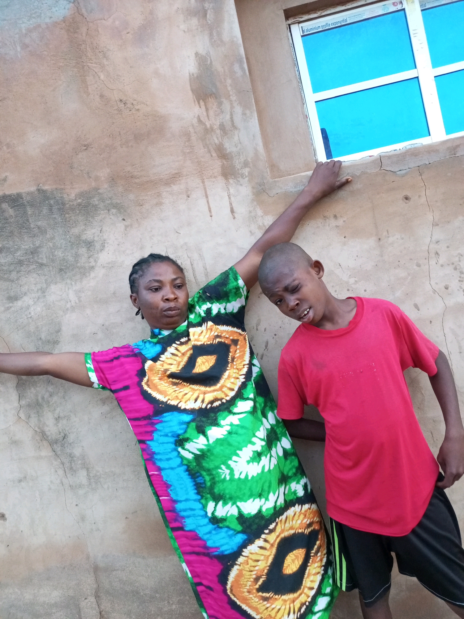 Two individuals, an adult woman and a young boy, are positioned in front of a textured, light brown, roughly plastered wall. The woman, wearing a vibrantly patterned dress with shades of pink, green, blue, and yellow, leans back with her left arm extended upward and to the left, touching the wall. Her hair is braided. The boy, to the woman's right, wears a red t-shirt and black shorts with green stripes. He is leaning forward, with his head tilted to his left and his left eye squinted, appearing to be winking. Behind the individuals, a window with a white frame and bright blue panes is visible. The overall scene suggests an outdoor setting. The provided context states the location as Tempure, Nigeria.