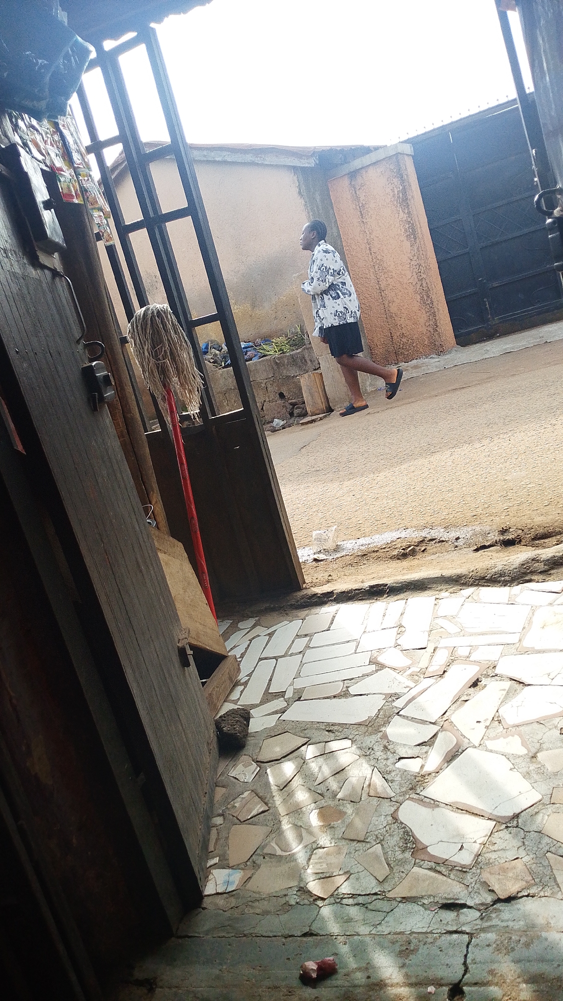 A person walks on a street in front of a building. The person is wearing a white shirt with black patterns and black shorts. The person is also wearing black sandals. The building has a large black gate. The street is paved with asphalt. The ground is covered with broken tiles. In the foreground, there is a wooden door and a mop. The lighting suggests it is daytime.