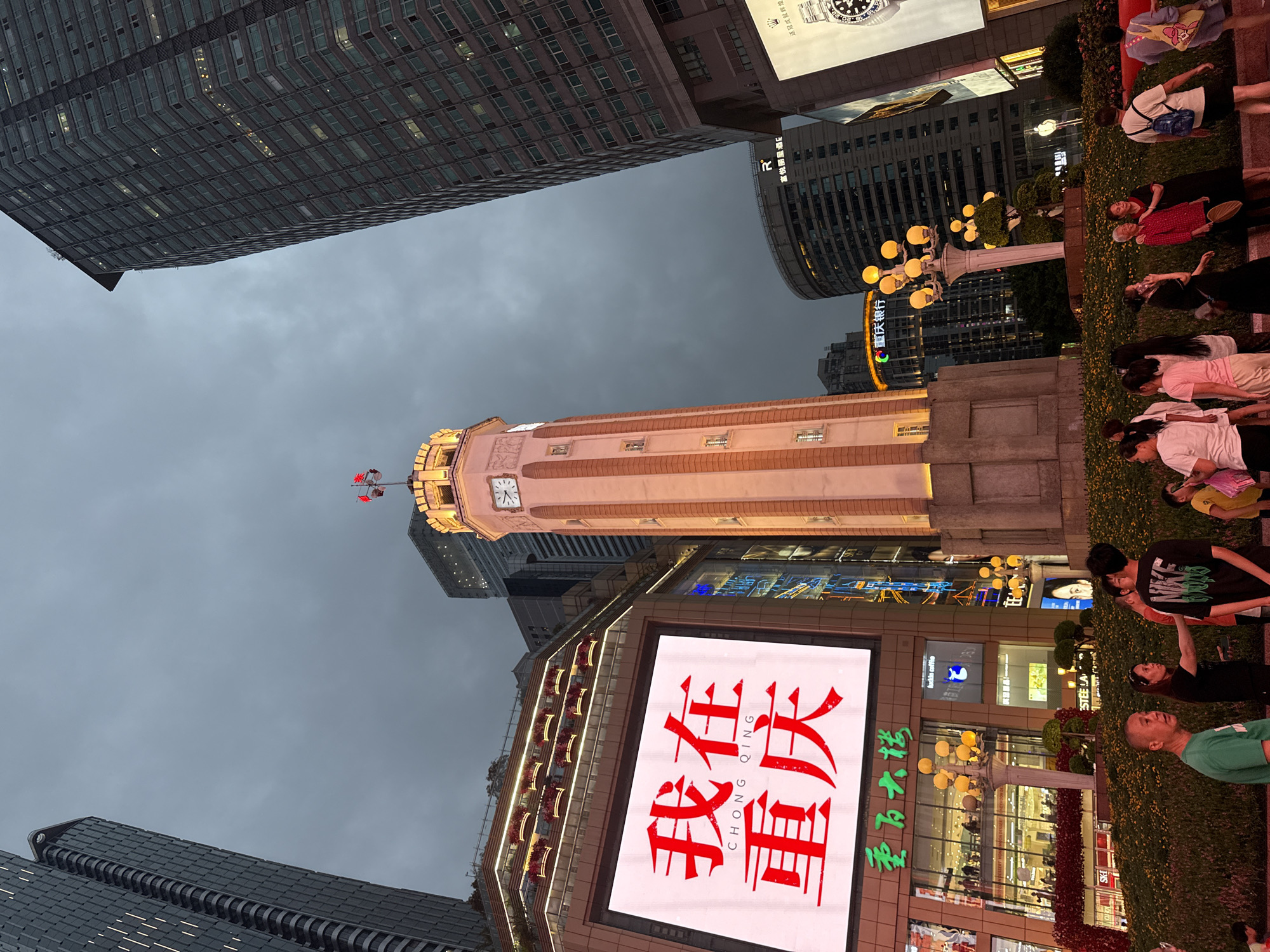 The image captures an evening scene in the Yuzhong District of Chongqing, China, showcasing a vibrant urban landscape. Dominating the center is a tall, illuminated clock tower with a red flag atop it. To the left, a large digital billboard displays "我在重庆" (I am in Chongqing) in bold red Chinese characters, situated above a multi-story building with numerous windows and decorative lighting. Surrounding the clock tower and billboard are modern skyscrapers, some with illuminated windows. In the foreground, a group of people are gathered, engaged in various activities, including conversations and strolling. The area is landscaped with bushes and small trees, and decorative streetlights with multiple globes illuminate the pathway. The sky is overcast, suggesting it is dusk or a cloudy evening.