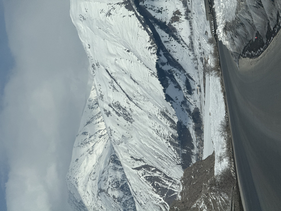 The image depicts a winter mountain landscape in the vicinity of Khurtisi, Georgia. A two-lane asphalt road, dark grey in color, curves from the lower-left foreground towards the right, with snow piled on its right shoulder and a metal guardrail partially obscured by snow. To the immediate left of the road, a steep, brown-grey rocky embankment with sparse, bare deciduous trees rises. The midground features a wide, snow-covered or frozen body of water, possibly a lake or river, stretching horizontally across a valley floor. On the far side of this frozen expanse, a small cluster of buildings constitutes a distant settlement, above which a dark, conical structure, possibly a tower or church, stands on a promontory at the base of the mountains. The background is dominated by multiple towering mountain ranges, all heavily blanketed in white snow. Exposed rock faces, dark patches of coniferous trees, and sparse deciduous vegetation are visible on the steep slopes. The sky above is partially overcast with grey clouds, particularly dense over the highest peaks, interspersed with areas of lighter blue sky. No individuals are visible. The overall environment indicates a cold, snowy, high-altitude setting.