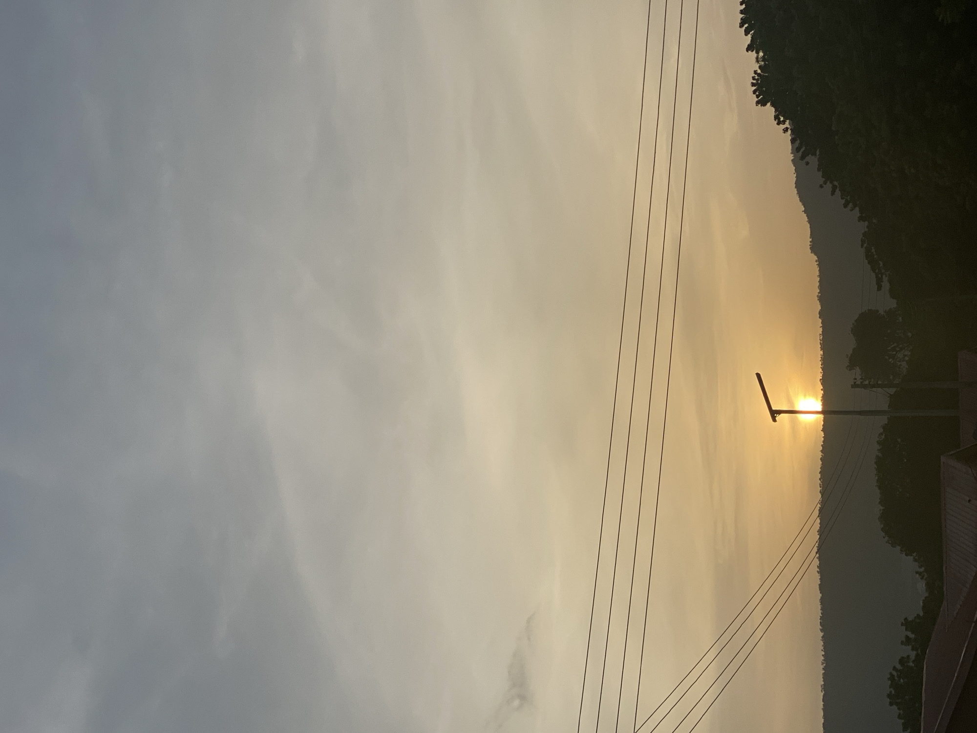 The image depicts a landscape at sunset in Adankolo, Nigeria. The sky transitions from greyish-blue on the left to golden-yellow towards the horizon, suggesting the setting sun. Silhouetted against the bright sky are the outlines of a tree line and a mountain ridge. Overhead power lines stretch across the frame, intersecting with a streetlight in the center. The light seems to be emanating directly from behind the hill. In the bottom right corner there appears to be a glimpse of a building.